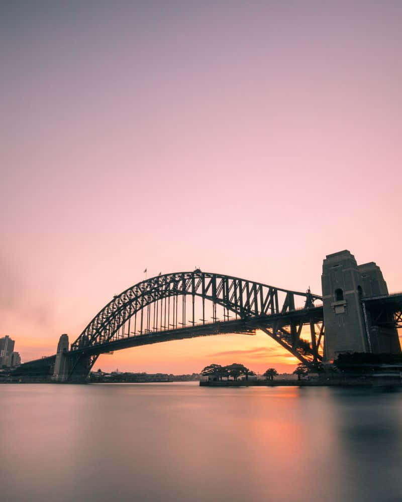 bridge 桌布 壁紙 — 雪梨港灣大橋在柔和粉紅與橙色的日落天空下呈現剪影，下方是平靜的水面。