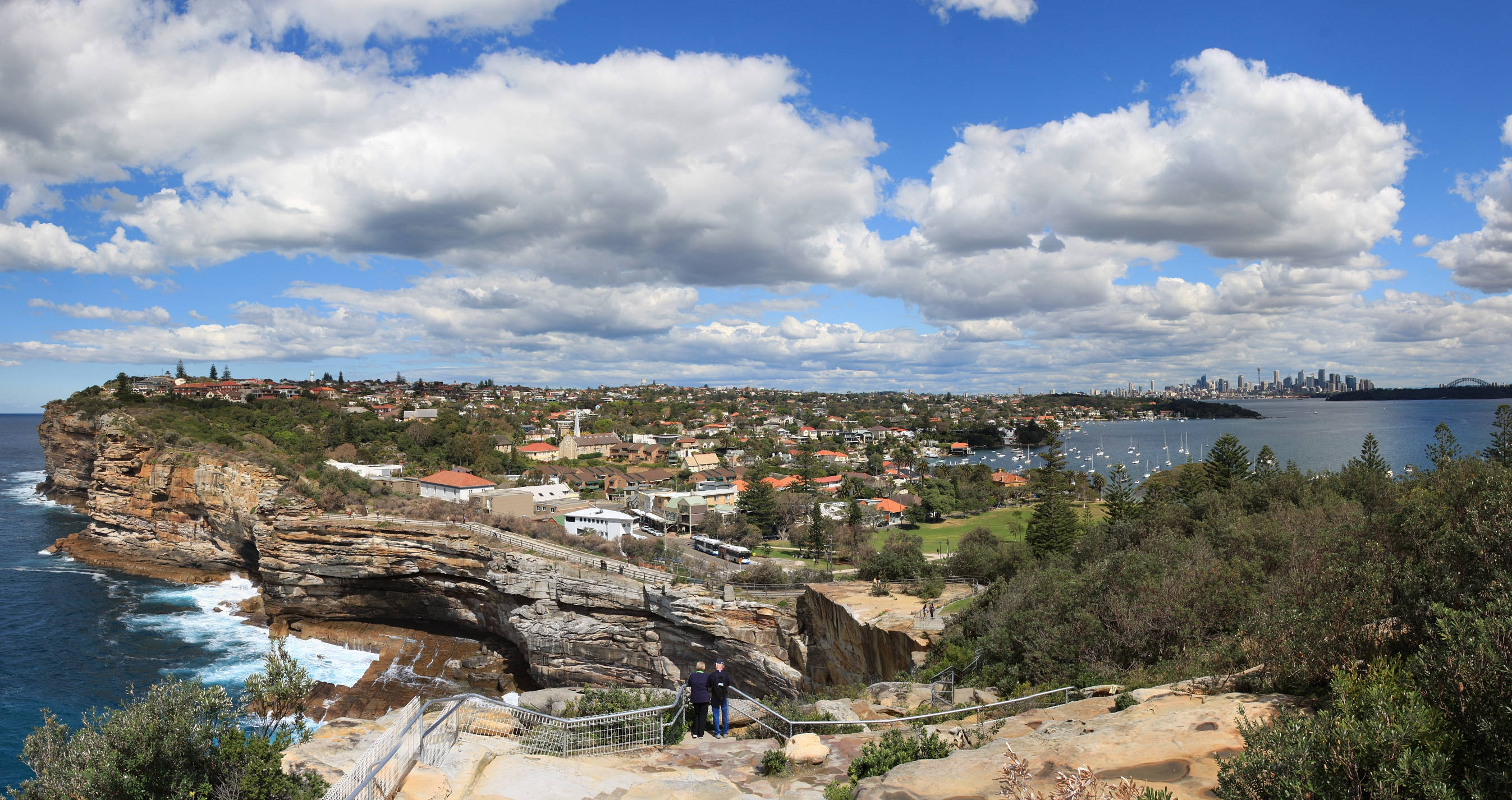 Sydney 桌布 壁紙 — 雪梨港的全景，懸崖、綠化和城市天際線在明亮的藍天和白雲下。