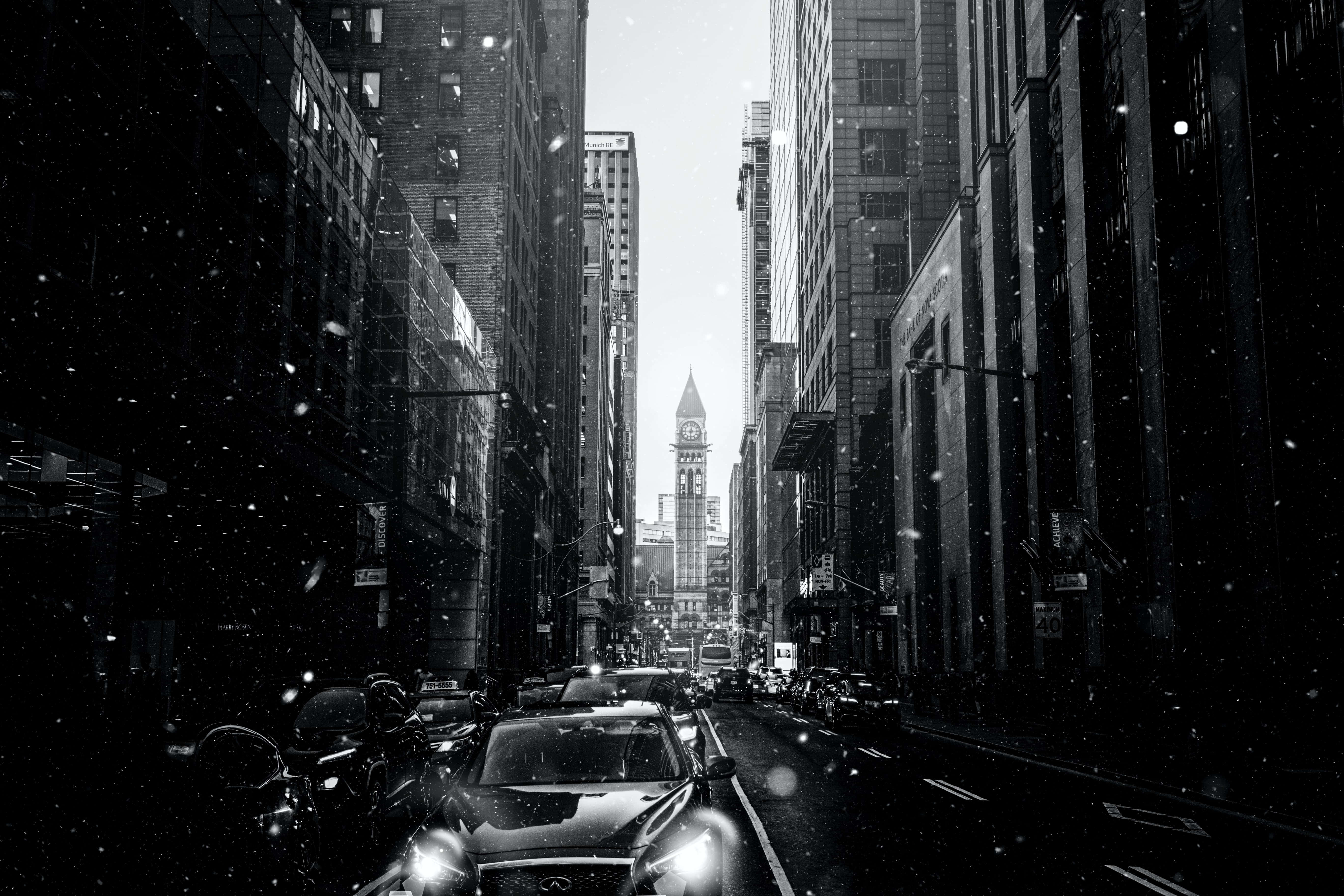 Aesthetic Black And White 桌布 壁紙 — 雪花在單色城市峽谷中飄蕩，車頭燈刺破黑暗，指向遠處的鐘樓。