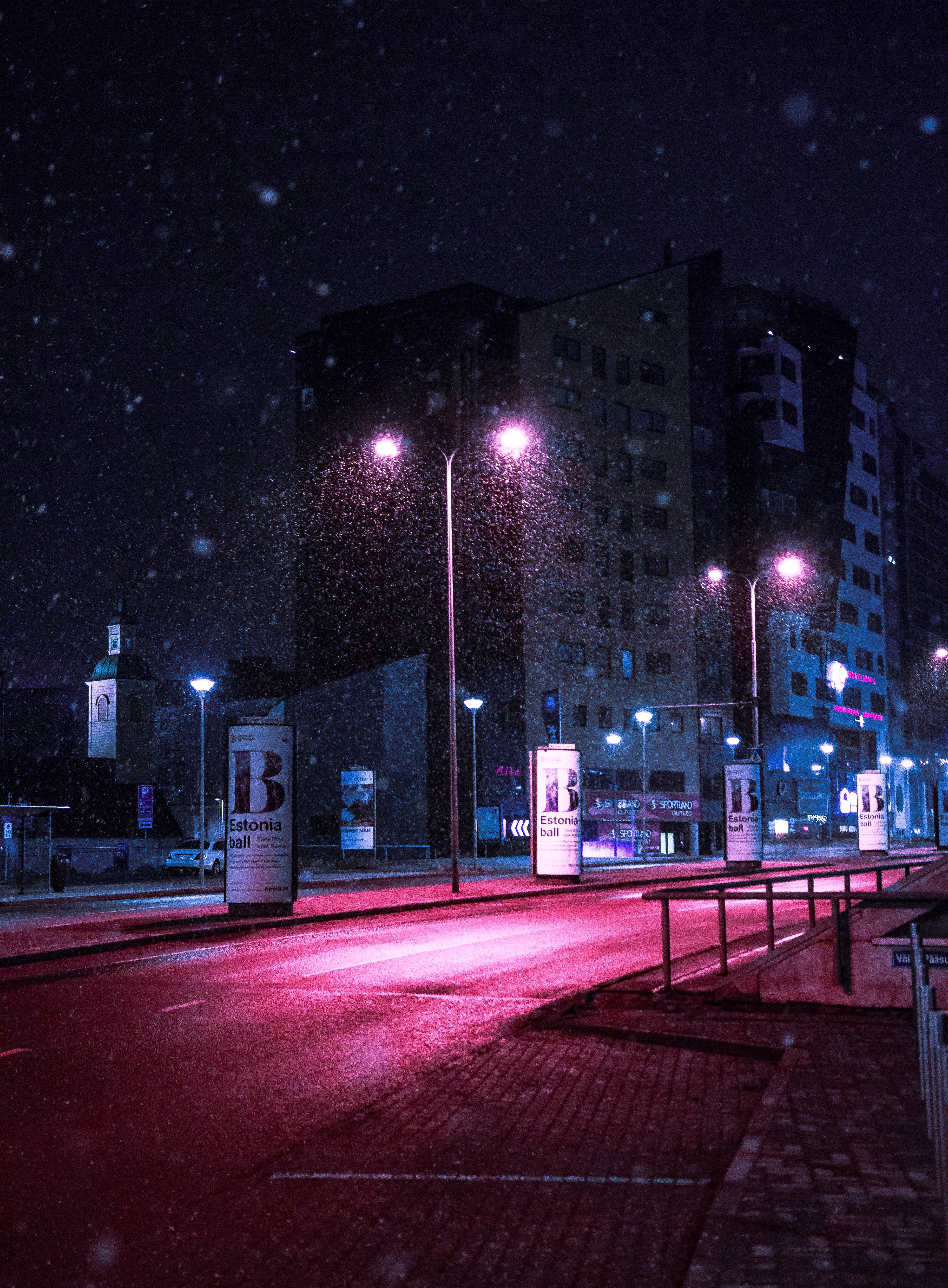 Road 桌布 壁紙 — 雪花在夜晚飄過霓虹燈照亮的城市街道，路燈在潮濕的路面和建築物上投下紅光。