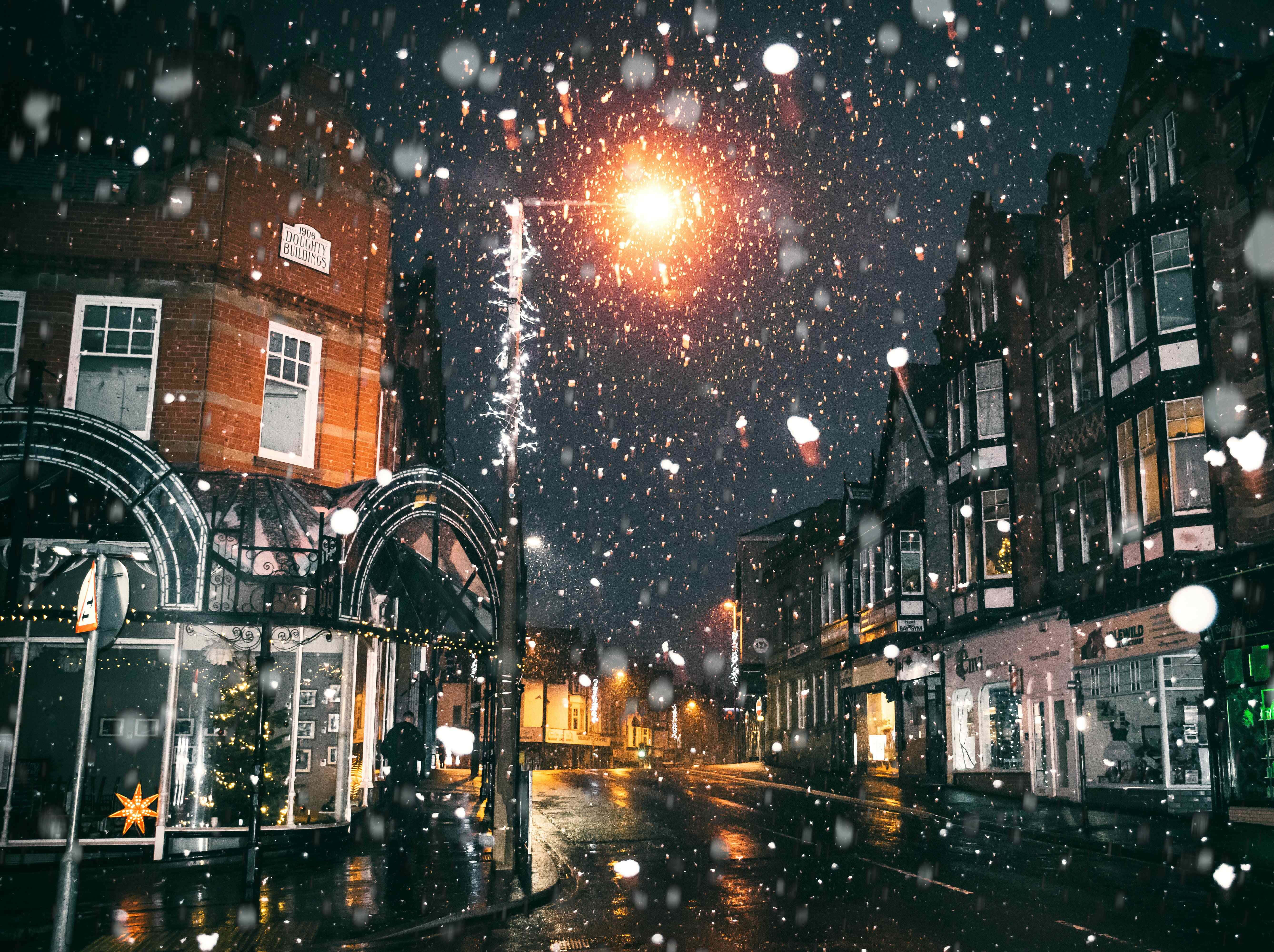 寒冷 桌布 壁紙 — 雪花在夜間飄過溫暖照亮的城市街道，潮濕的路面反射著發光的街燈和節日櫥窗。
