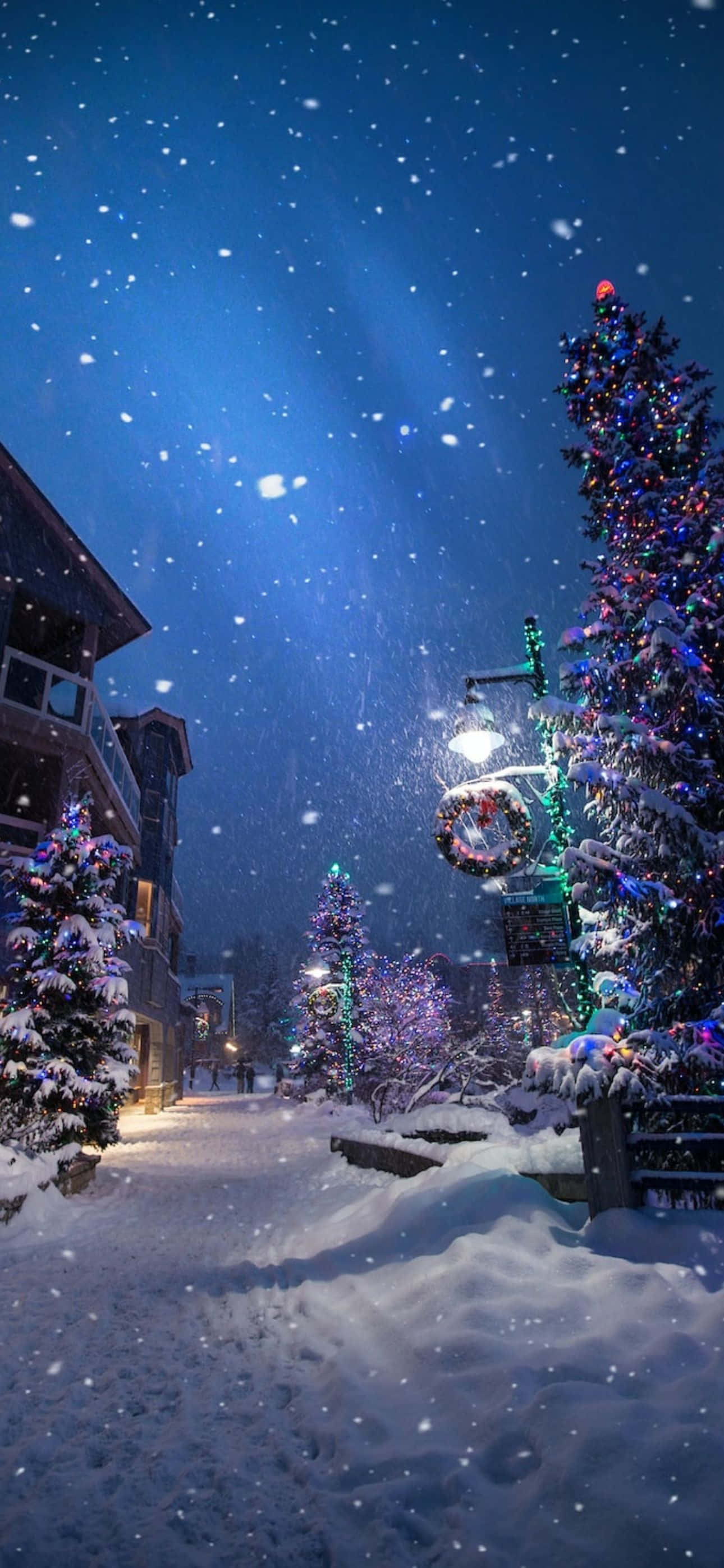 聖誕節 iPhone 桌布 壁紙 — 雪花在星光閃爍的夜晚飄落，節日燈飾在白雪覆蓋的樹木和建築物上閃爍，營造出一個舒適的冬季村莊。