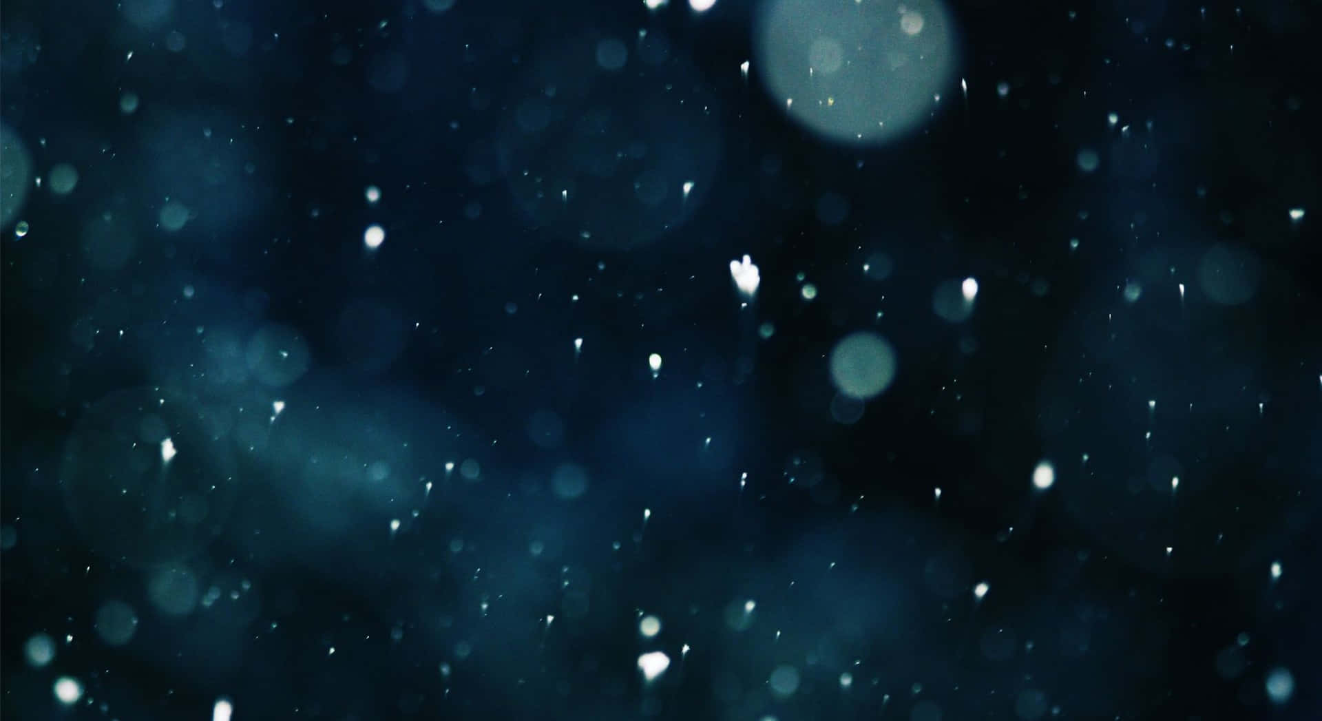 Cool Dark Desktop 桌布 壁紙 — 雪花在深藍色散景背景上輕輕飄落，伴隨著發光球體和柔和的光粒子。