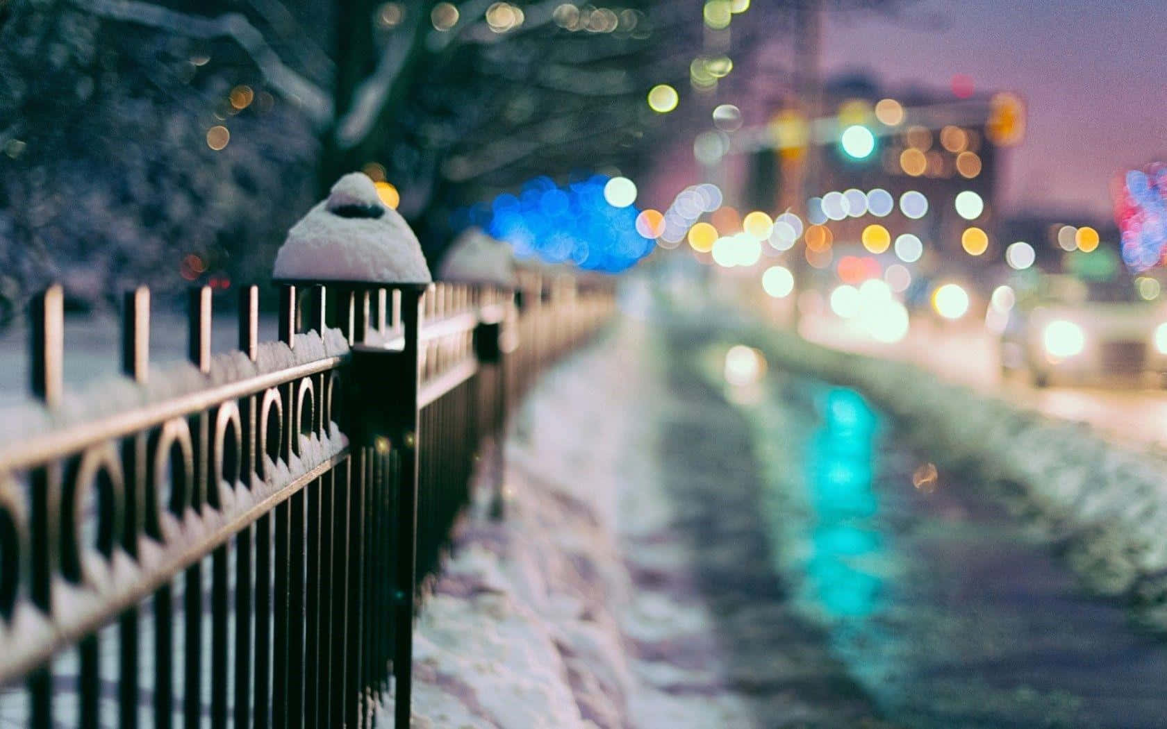 Winter Cityscape 桌布 壁紙 — 雪花輕灑在黑色鐵欄杆上，旁邊濕漉漉的人行道倒映著夜晚的城市燈光。