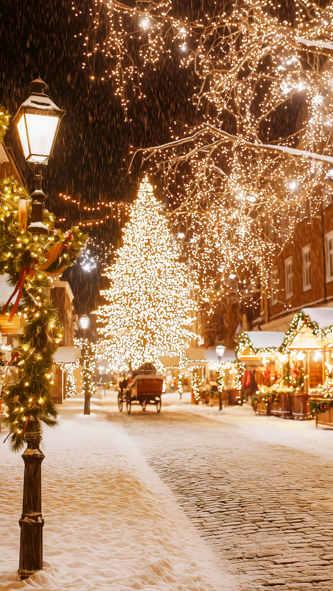 Christmas Wallpapers 壁紙 — 雪花輕輕飄落在鵝卵石街道上，街道兩旁掛滿了發光的燈串和高聳的聖誕樹。
