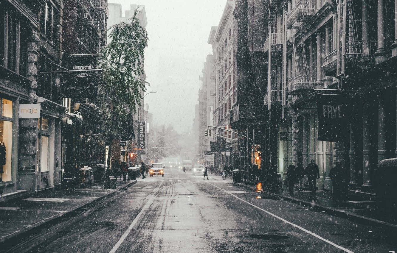 Icy Road 桌布 壁紙 — 雪花飄過一條寧靜的城市街道，兩旁是歷史建築和發光的商店櫥窗，天空灰濛濛。