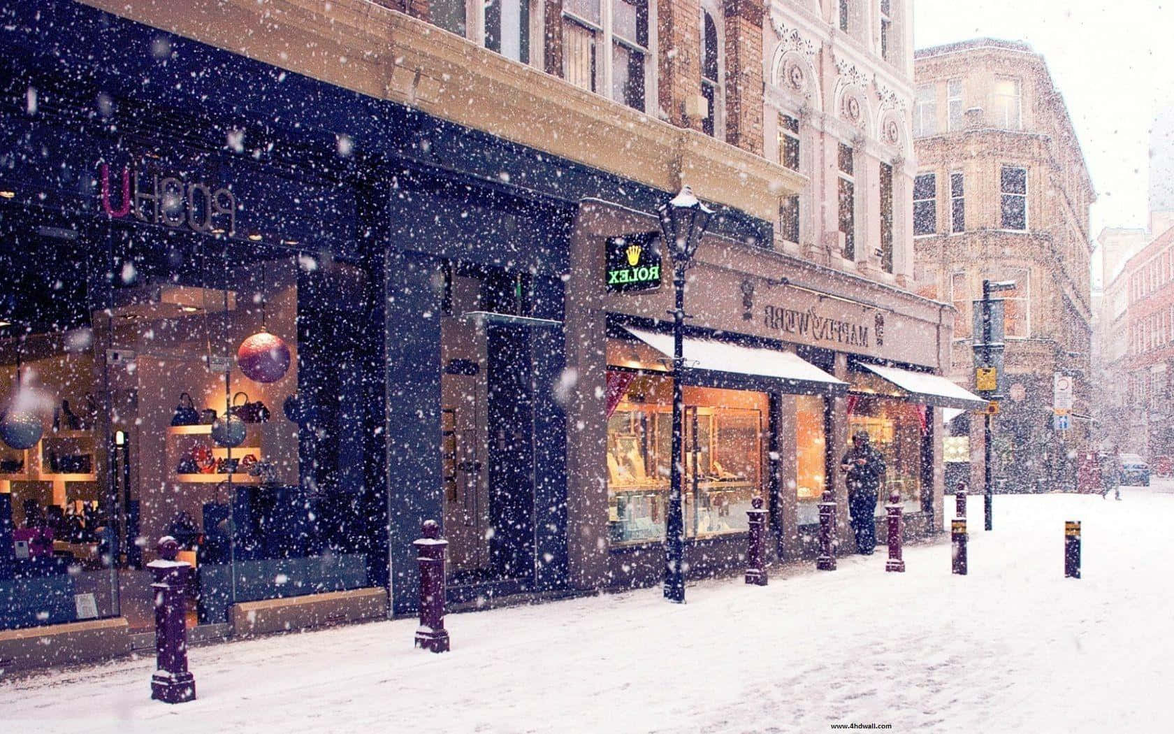 Winter Cityscape 桌布 壁紙 — 雪花飄過一條寧靜的城市街道，兩旁是溫暖燈光的商店和歷史悠久的磚砌建築。