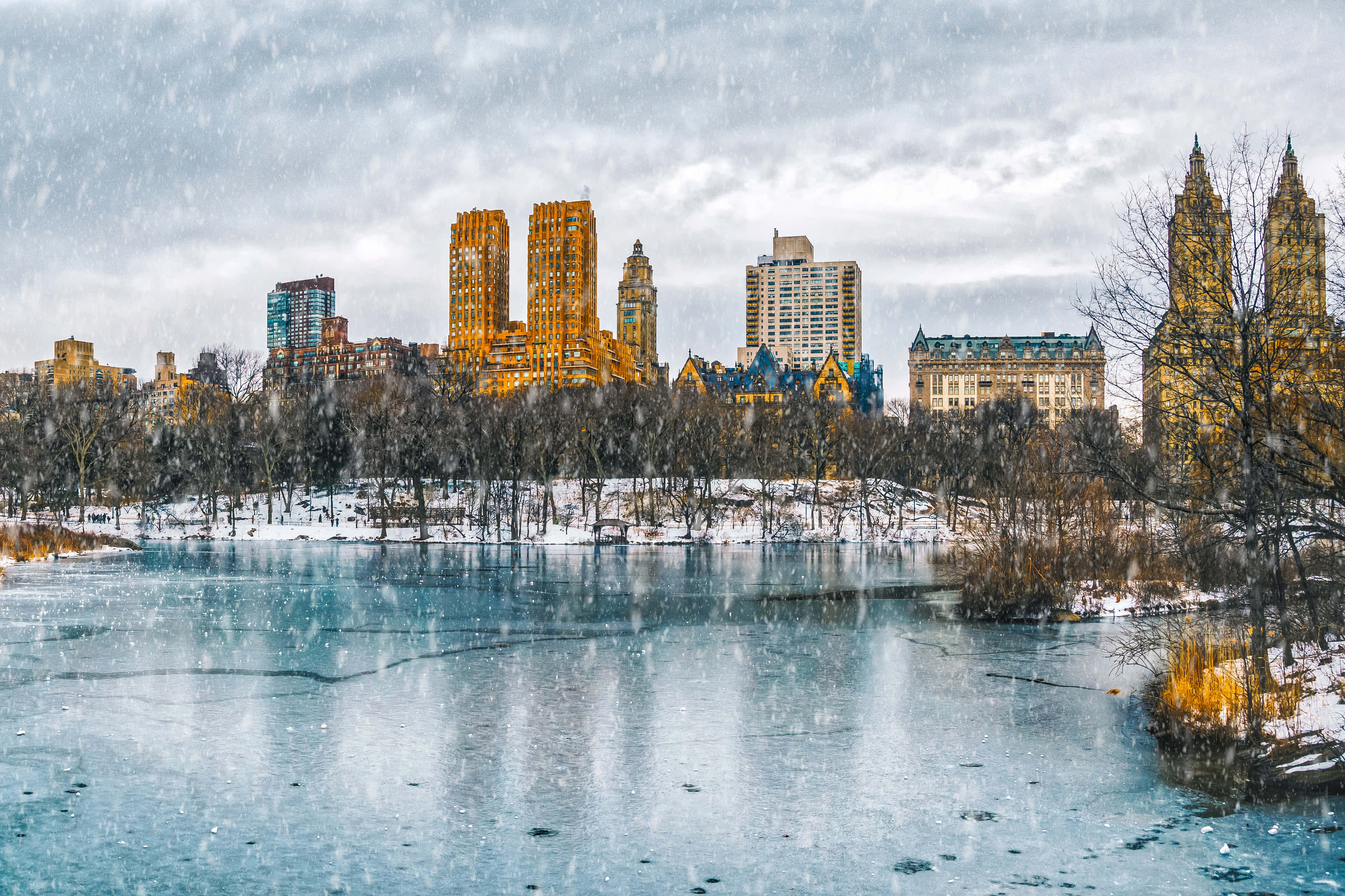 聖誕節 桌布 壁紙 — 雪落在中央公園結冰的湖面上，城市摩天大樓在背景中溫暖地閃耀。