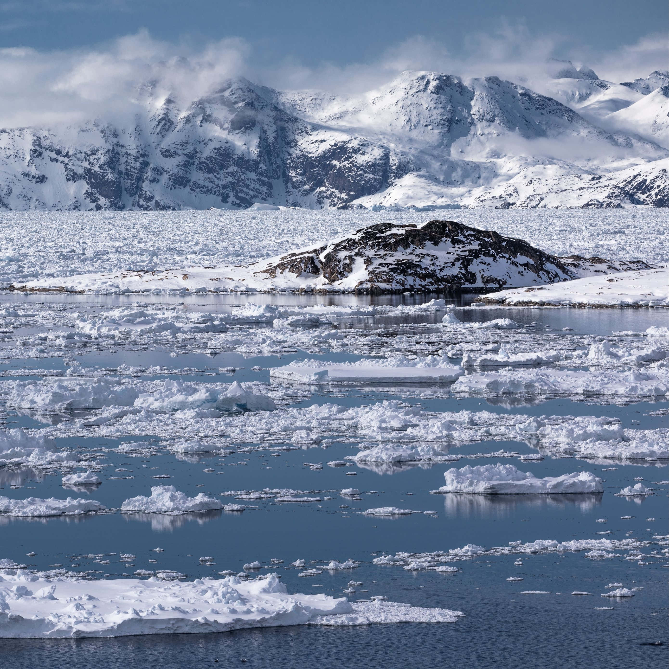 Greenland 桌布 壁紙 — 雪覆蓋的山脈在冰封的海面上聳立，海面上散佈著浮冰和岩石露頭。