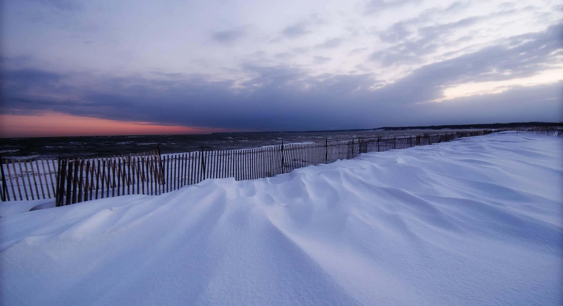 Winter Beach 桌布 壁紙 — 雪覆蓋的沙丘上有一道木柵欄，在紫色和粉紅色的天空下延伸向暮色海洋。