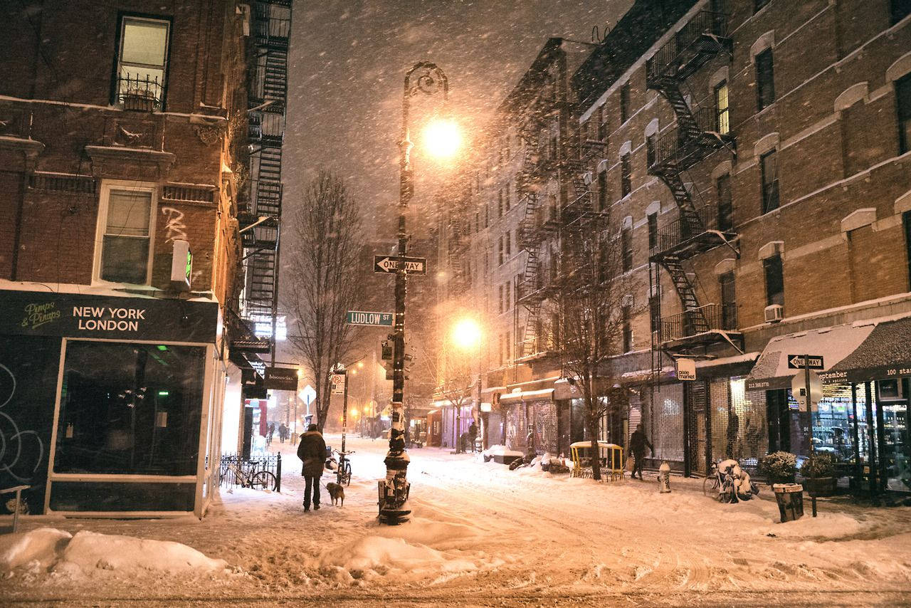 雪 桌布 壁紙 — 雪覆蓋著寧靜的城市街道，溫暖的路燈在磚砌建築和遛狗的行人身上投下金色的光芒。