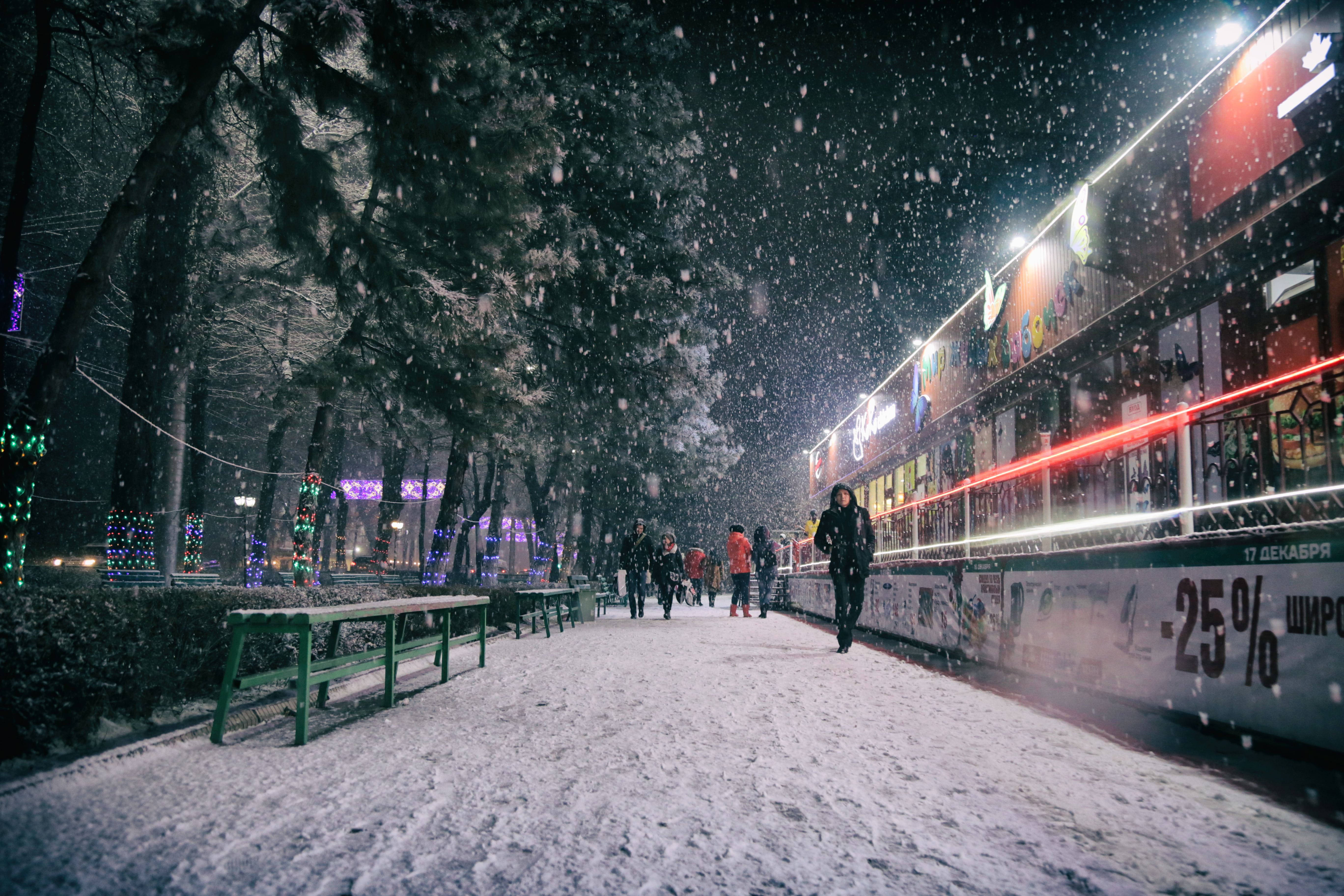 Temperature 桌布 壁紙 — 雪輕輕落在夜間的城市人行道上，兩旁是閃爍的節日燈飾和積雪的樹木。