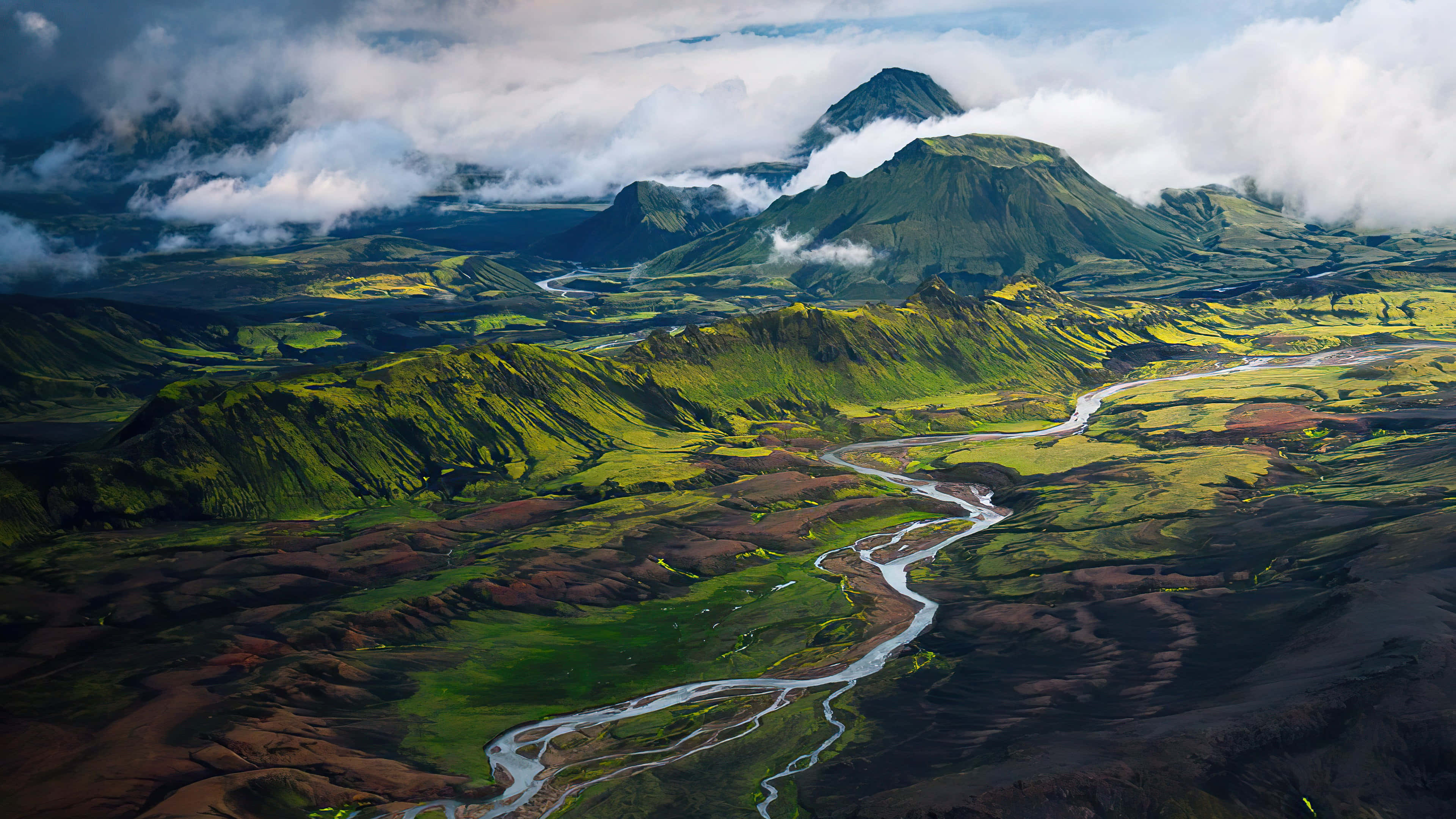 Iceland Desktop 桌布 壁紙 — 雲層覆蓋的山脈下，蜿蜒的河流穿過充滿活力的綠色和棕色火山山谷的鳥瞰圖。