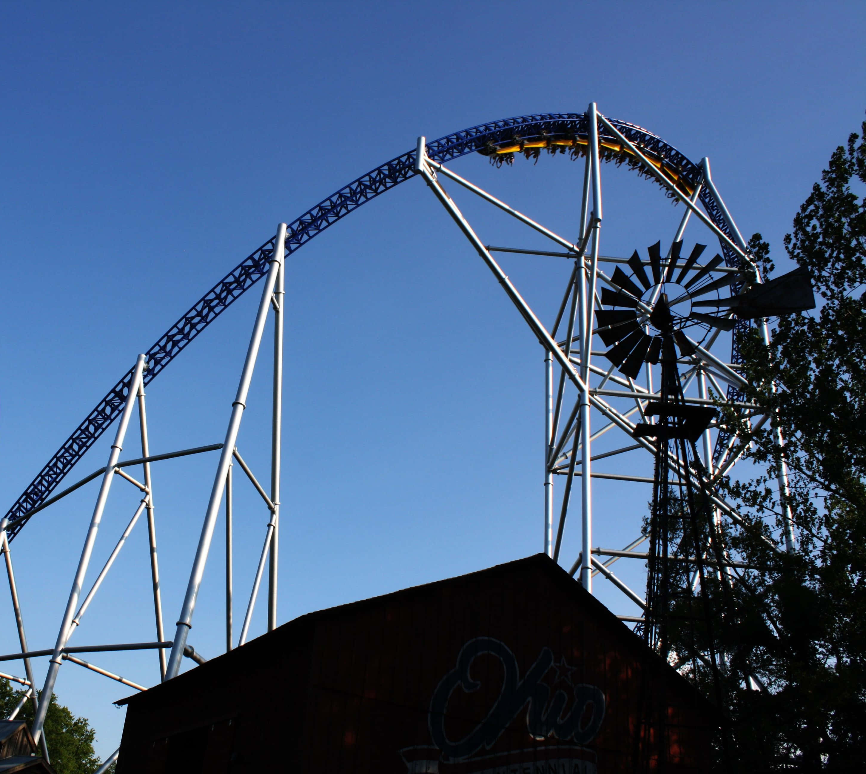 Roller Coaster 桌布 壁紙 — 雲霄飛車在晴朗的藍天下飛馳，下方有風車和質樸的建築。