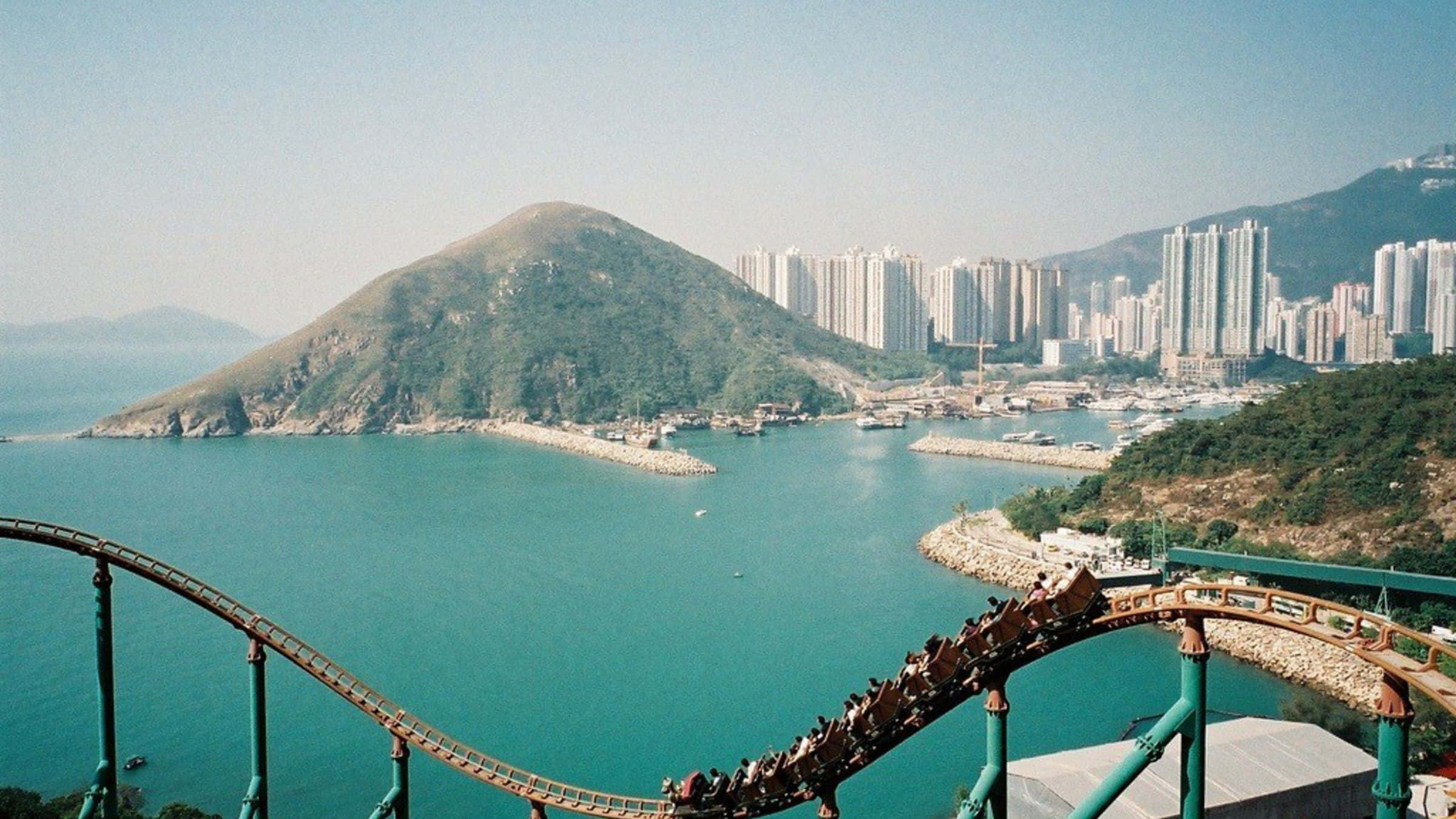 Roller Coaster 桌布 壁紙 — 雲霄飛車在碧綠色的水上彎曲，背景有綠色山丘和城市天際線。