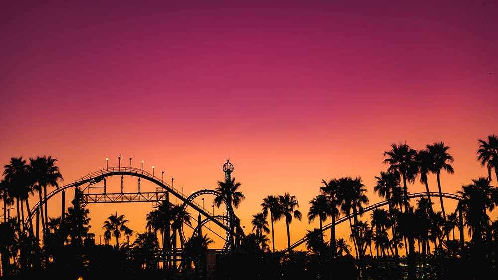 Roller Coaster 桌布 壁紙 — 雲霄飛車在鮮豔的粉紅色和橙色日落天空下剪影，下方有棕櫚樹。
