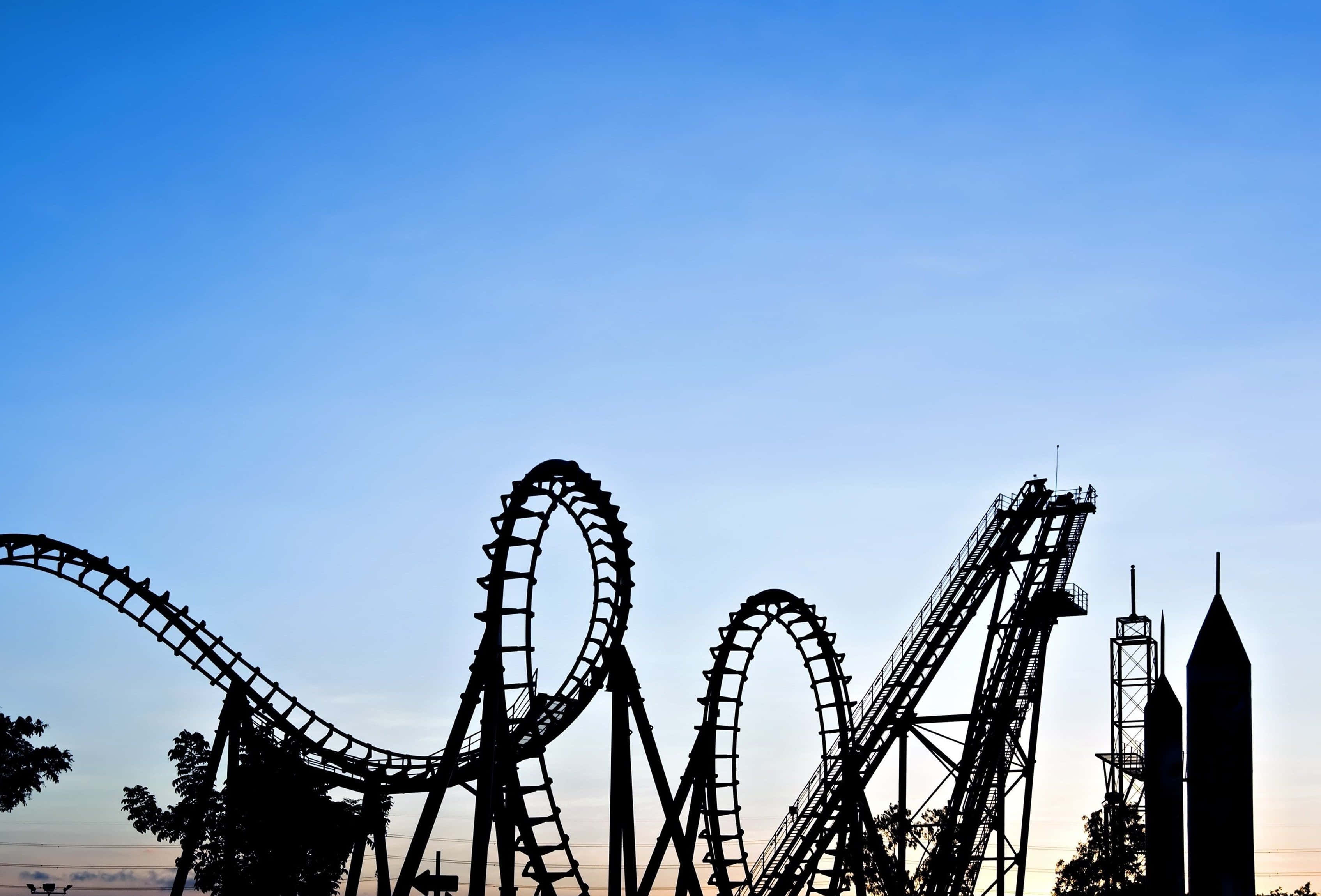 Roller Coaster 桌布 壁紙 — 雲霄飛車的迴圈和塔樓在黃昏時分以剪影形式映襯著漸變的藍色天空。