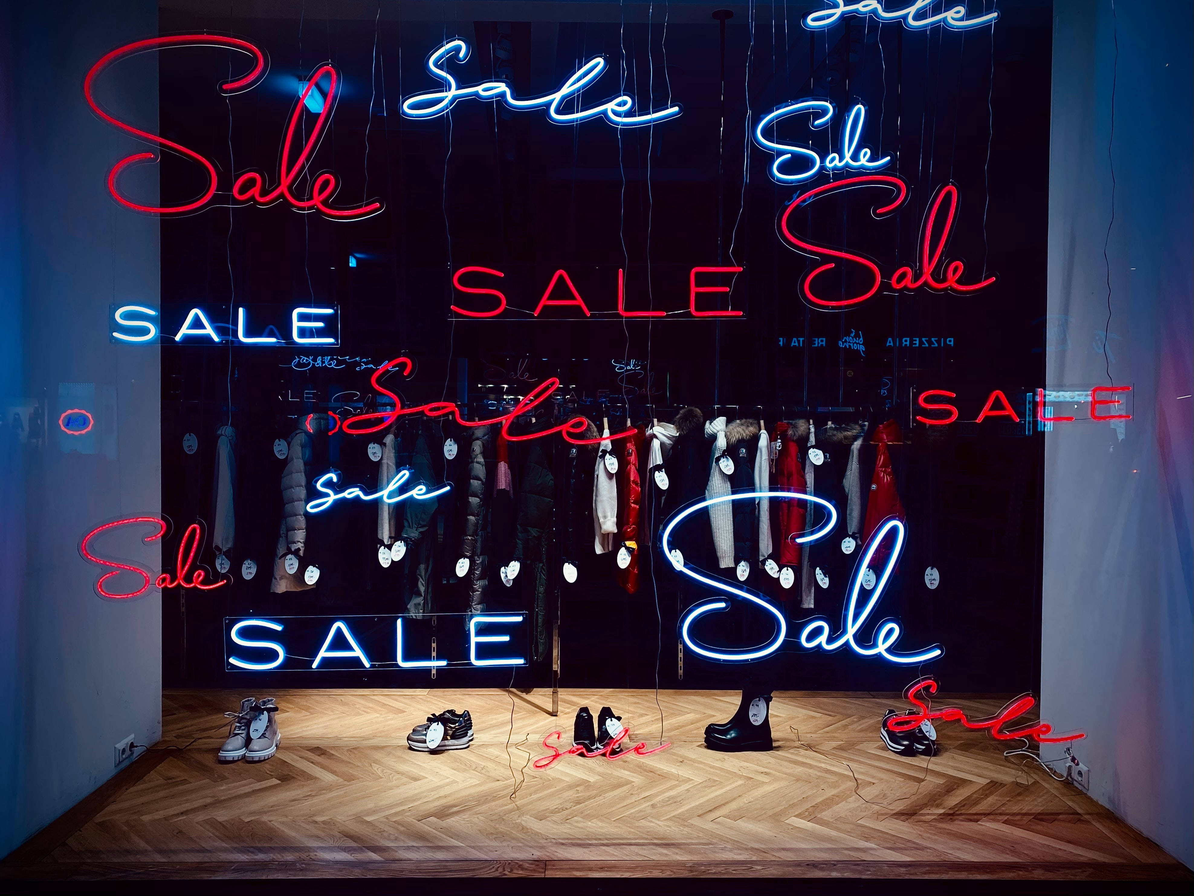 Sales 桌布 壁紙 — 零售店櫥窗展示中，發光的紅色和藍色霓虹燈「Sale」標誌懸掛在木地板上方的衣服和鞋子架上。
