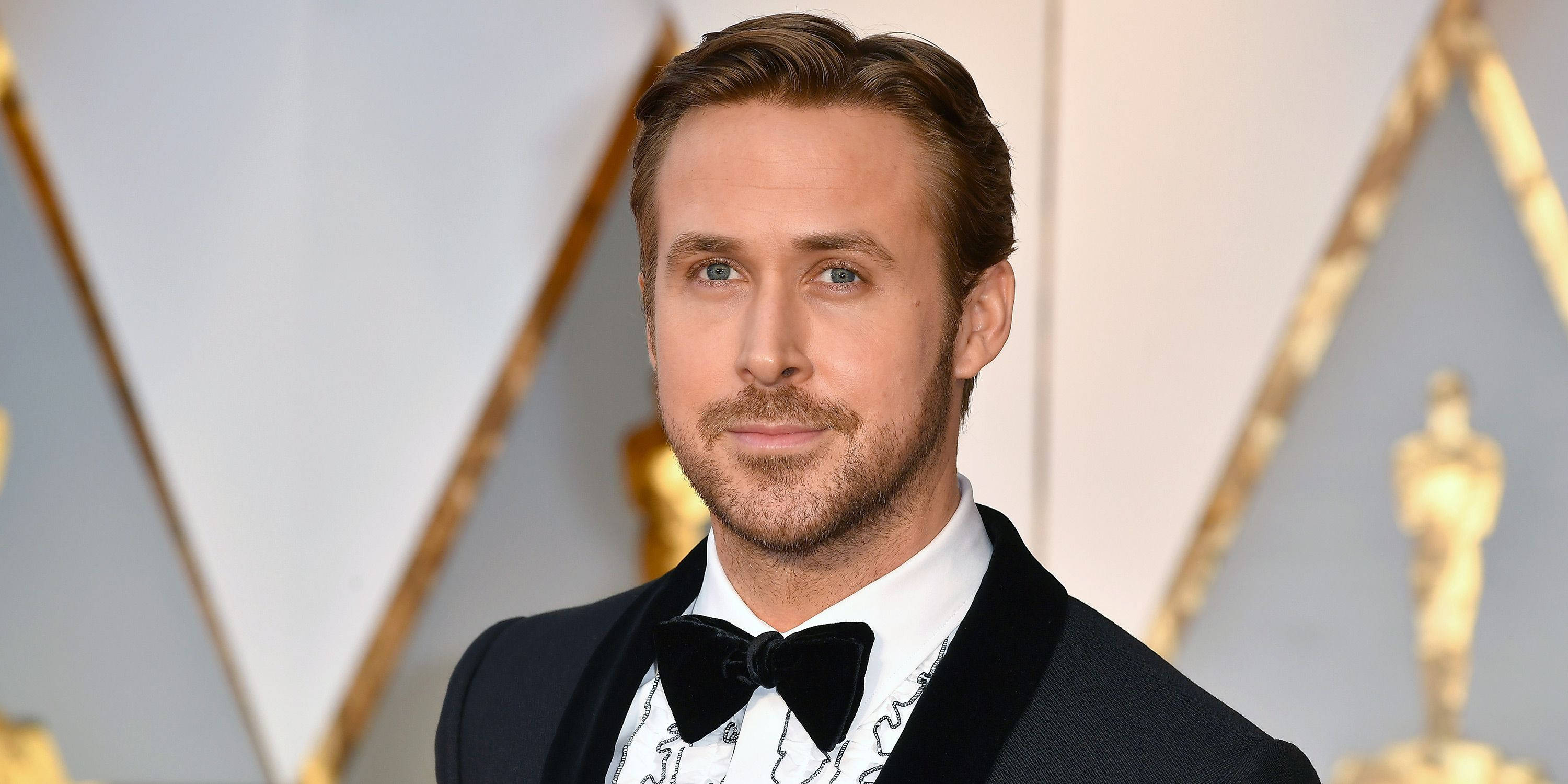 Ryan Gosling 桌布 壁紙 — 雷恩葛斯林身穿黑色燕尾服，搭配天鵝絨翻領和蝴蝶結，以金色奧斯卡獎座和幾何金色圖案為背景擺姿勢。