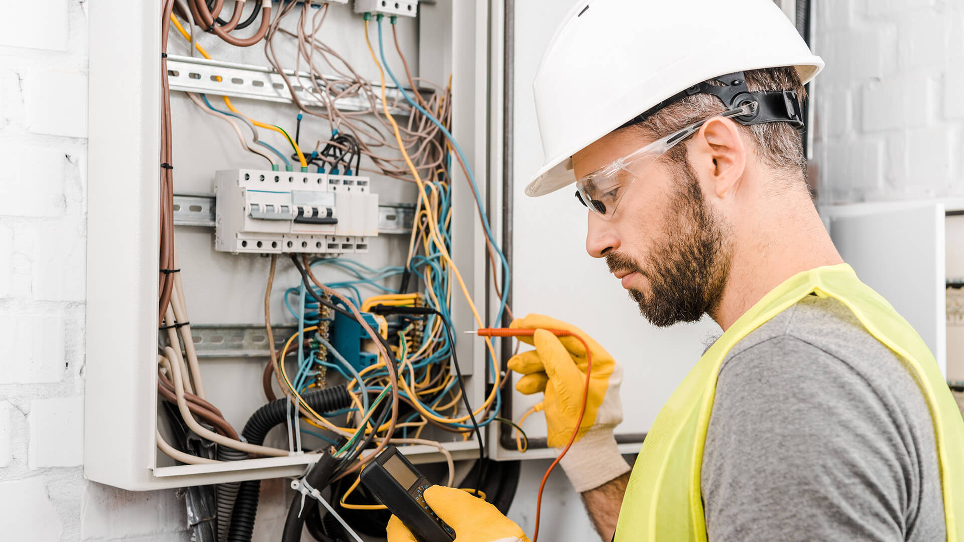 professional 桌布 壁紙 — 電工戴著頭盔和安全背心，用萬用電錶測試打開的電箱內的接線。