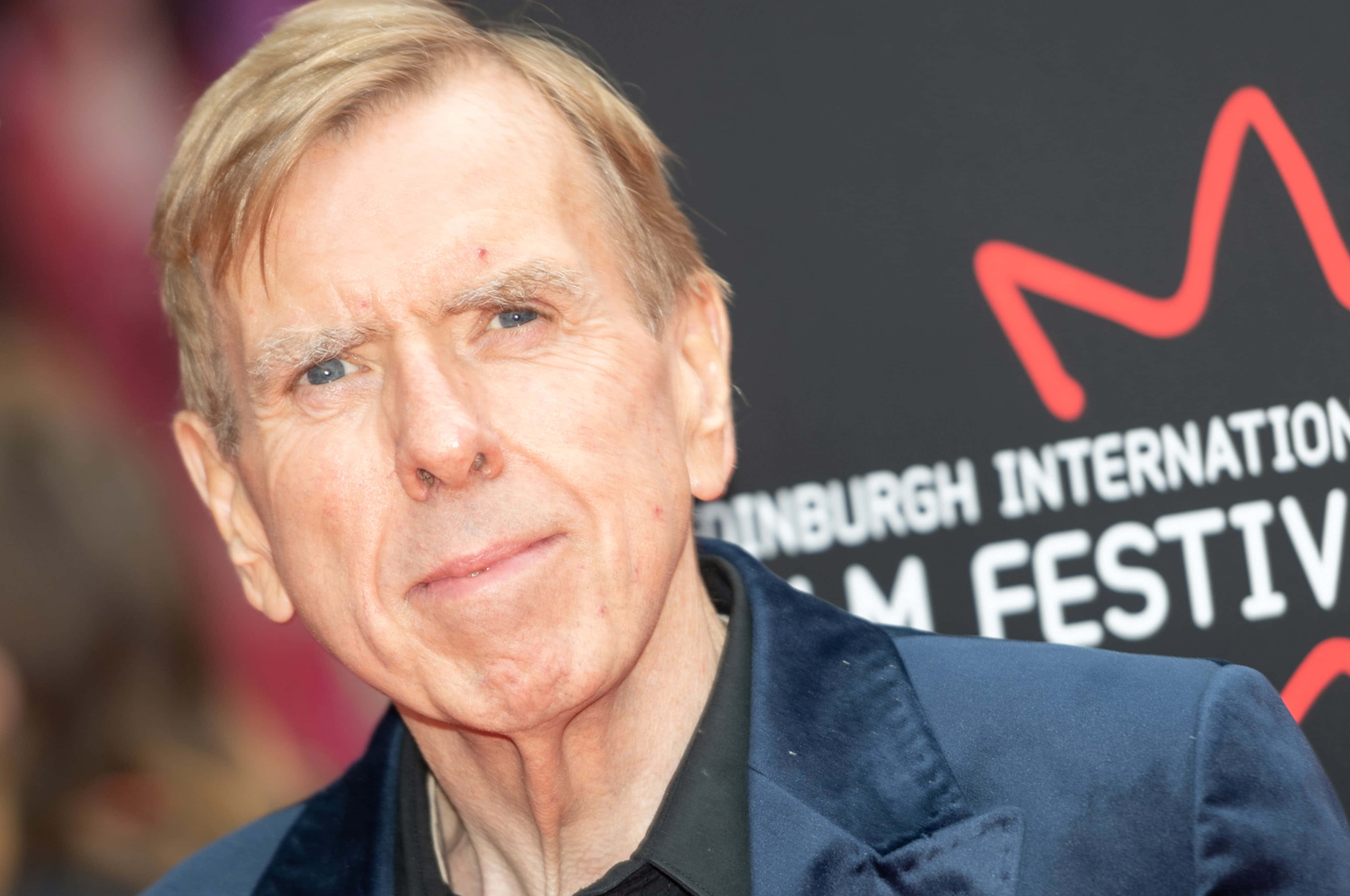 Timothy Spall 桌布 壁紙 — 電影節活動中，一名金髮藍眼男子穿著深藍色天鵝絨外套的特寫肖像。