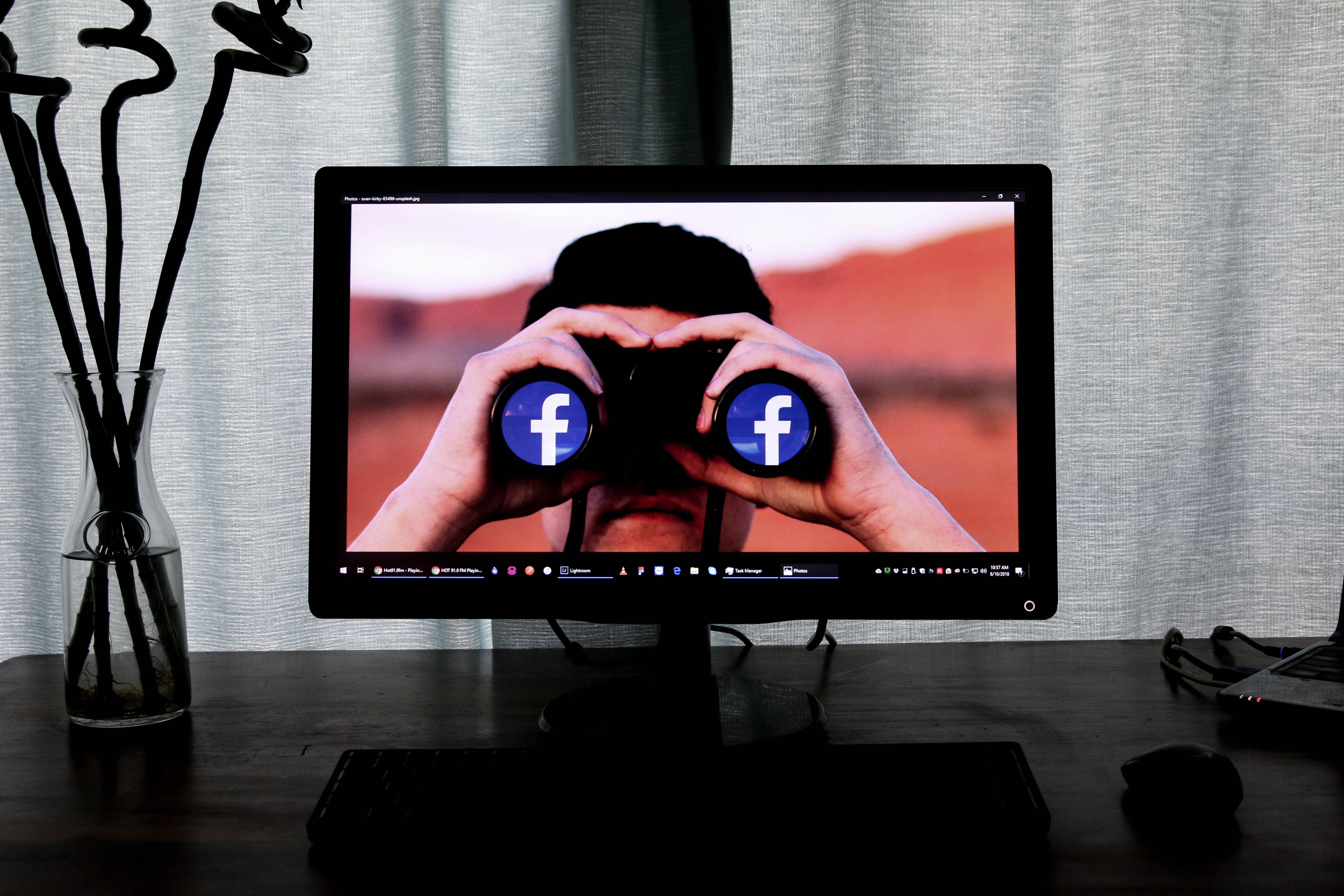Facebook 桌布 壁紙 — 電腦顯示器顯示一個人使用雙筒望遠鏡，鏡片上有Facebook標誌，背景是沙漠，放在一張深色桌子上，旁邊有一個插著黑色樹枝的花瓶。