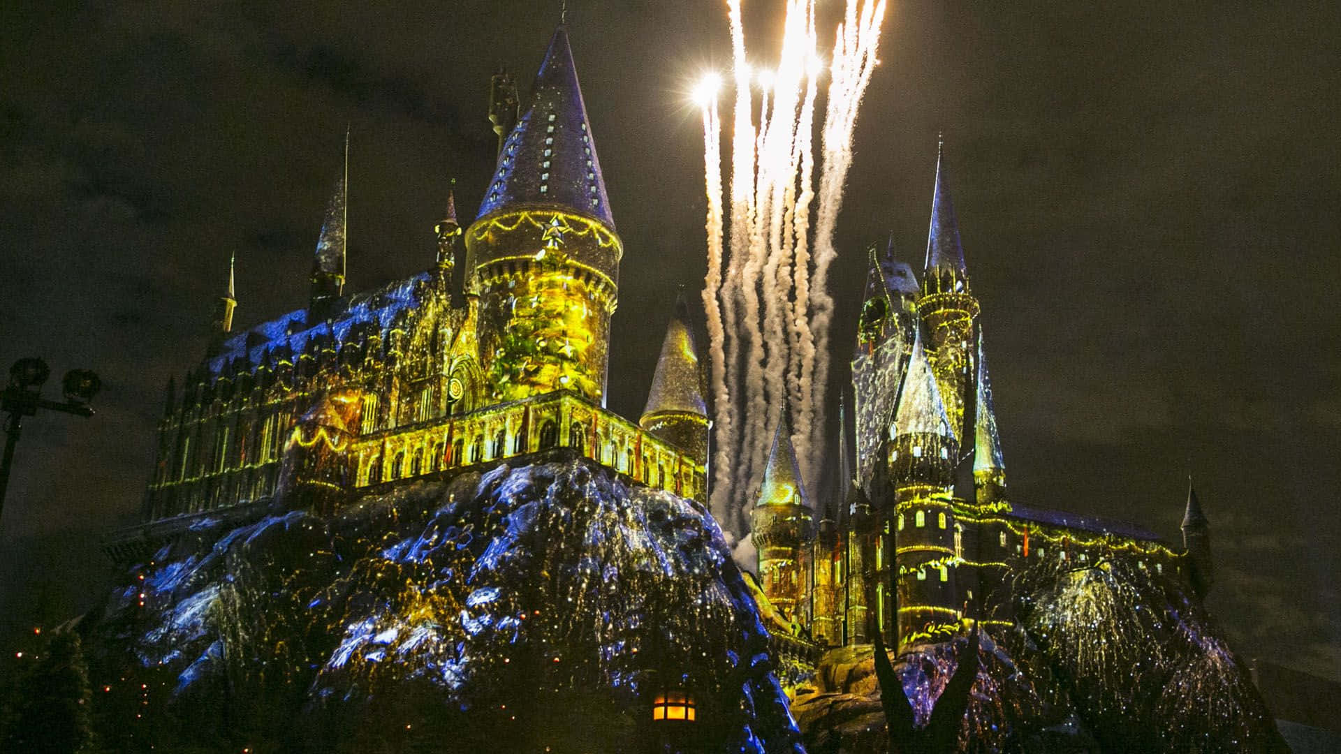 Harry Potter Christmas 桌布 壁紙 — 霍格華茲城堡在夜晚被金色燈光和尖塔上方爆炸的煙火照亮。
