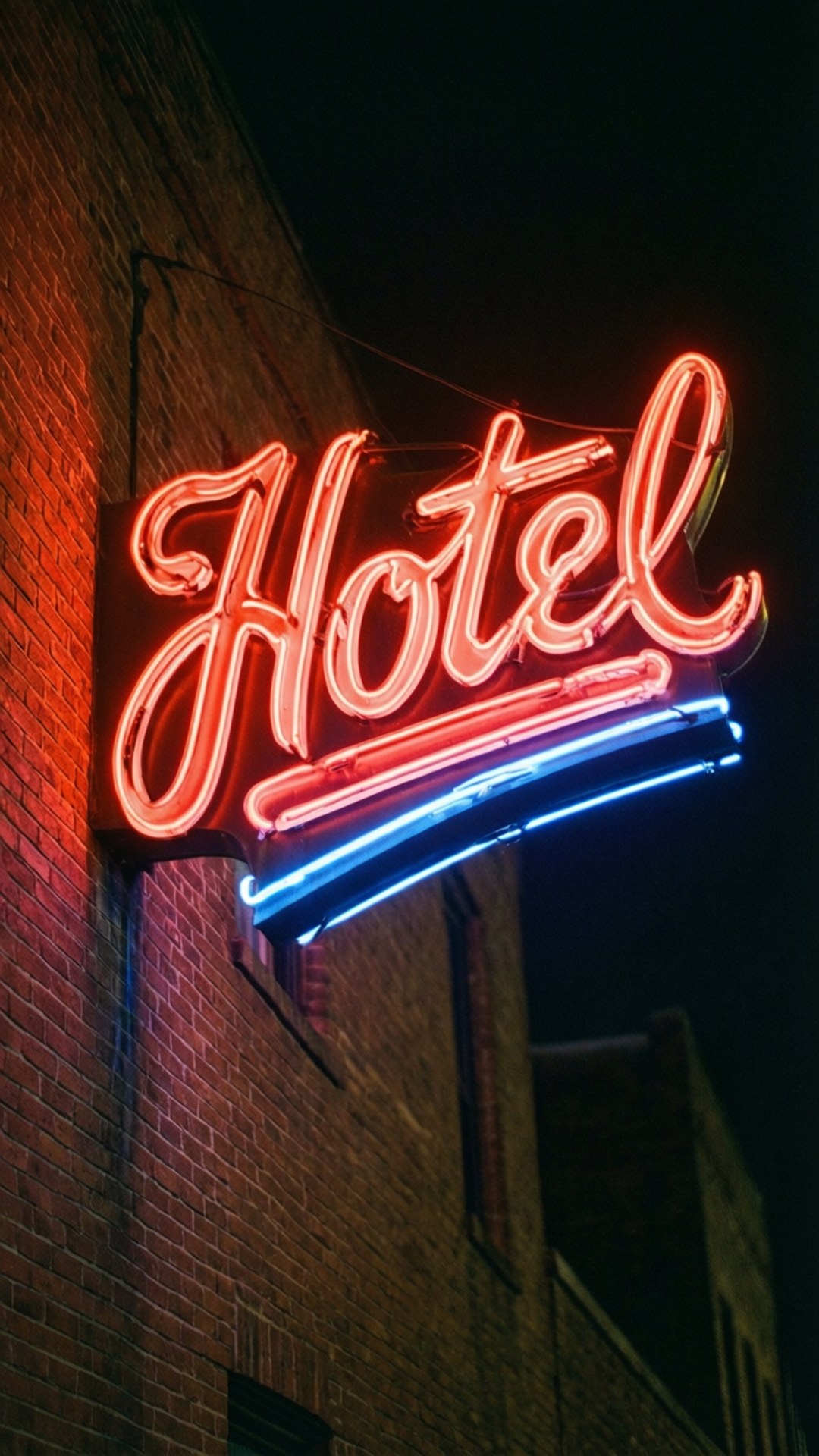 霓虹旅館招牌 壁紙 — 一個發光的紅藍霓虹燈招牌，拼寫著'Hotel'，照亮了磚砌建築，映襯著黑暗的夜空。