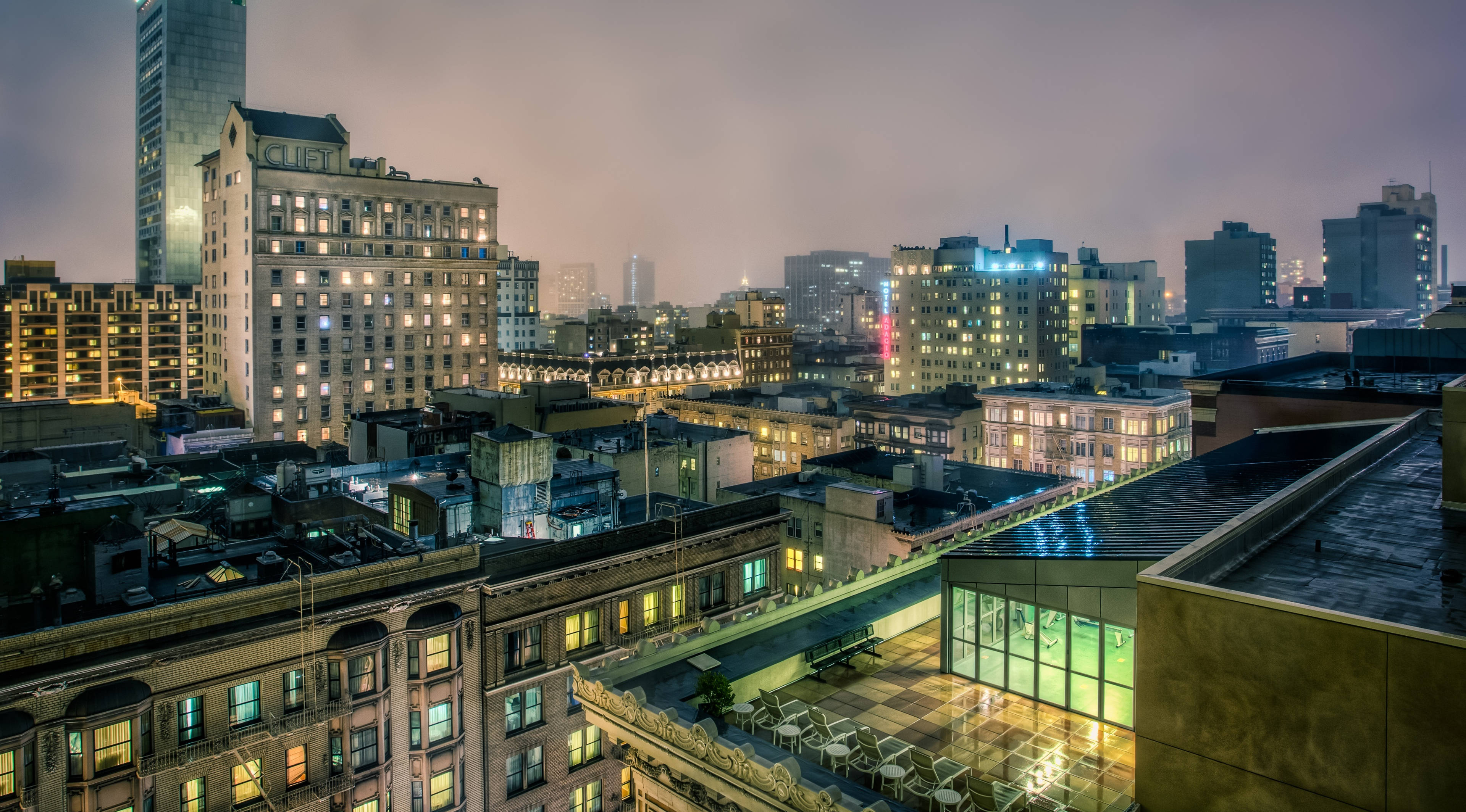 View 桌布 壁紙 — 霧氣瀰漫的城市夜景，建築物燈火通明，屋頂露台在柔和暮色中閃爍微光。