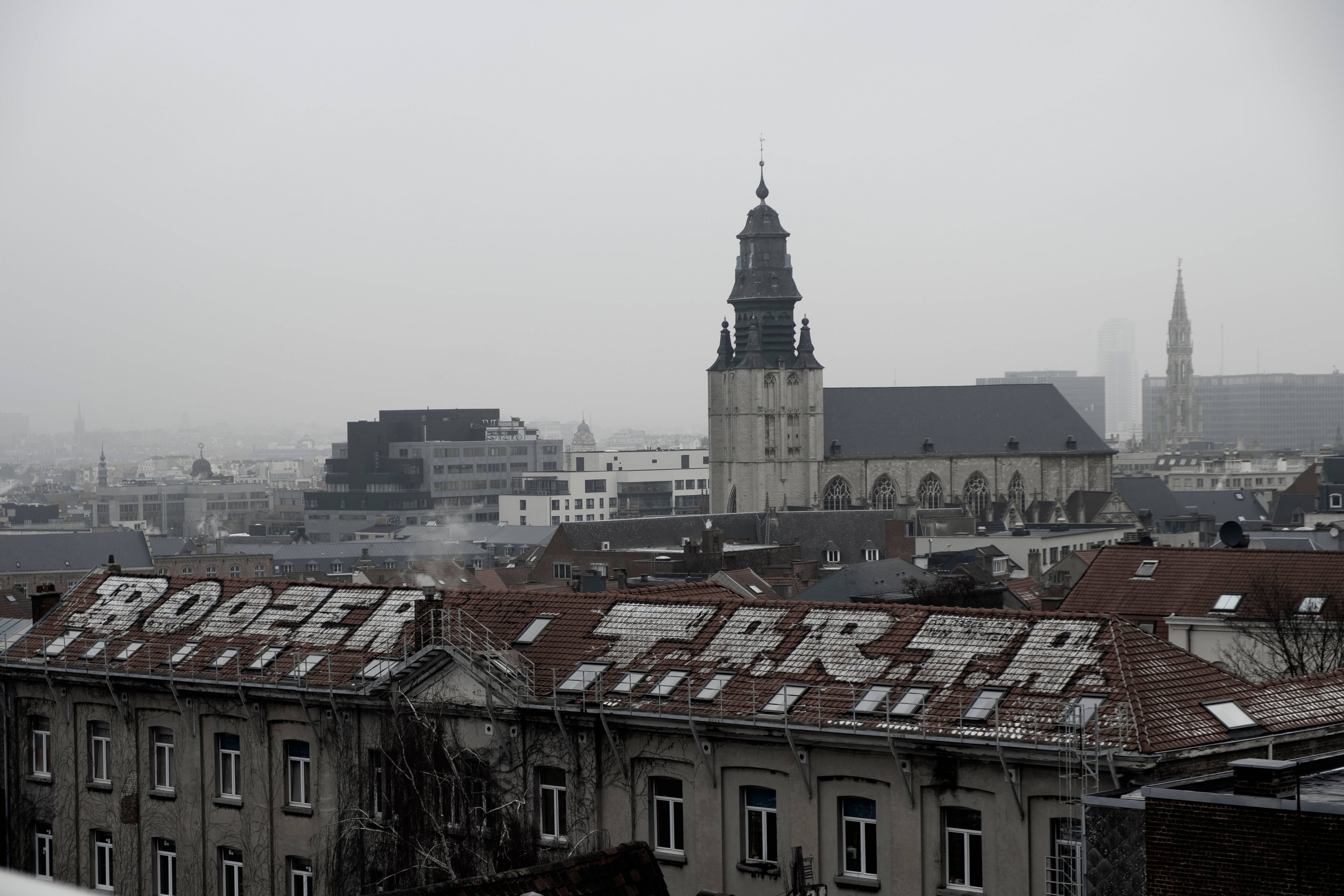 Belgium 桌布 壁紙 — 霧氣瀰漫的城市天際線，以一座歷史悠久的教堂塔樓為主，聳立於帶有褪色塗鴉的屋頂之上。