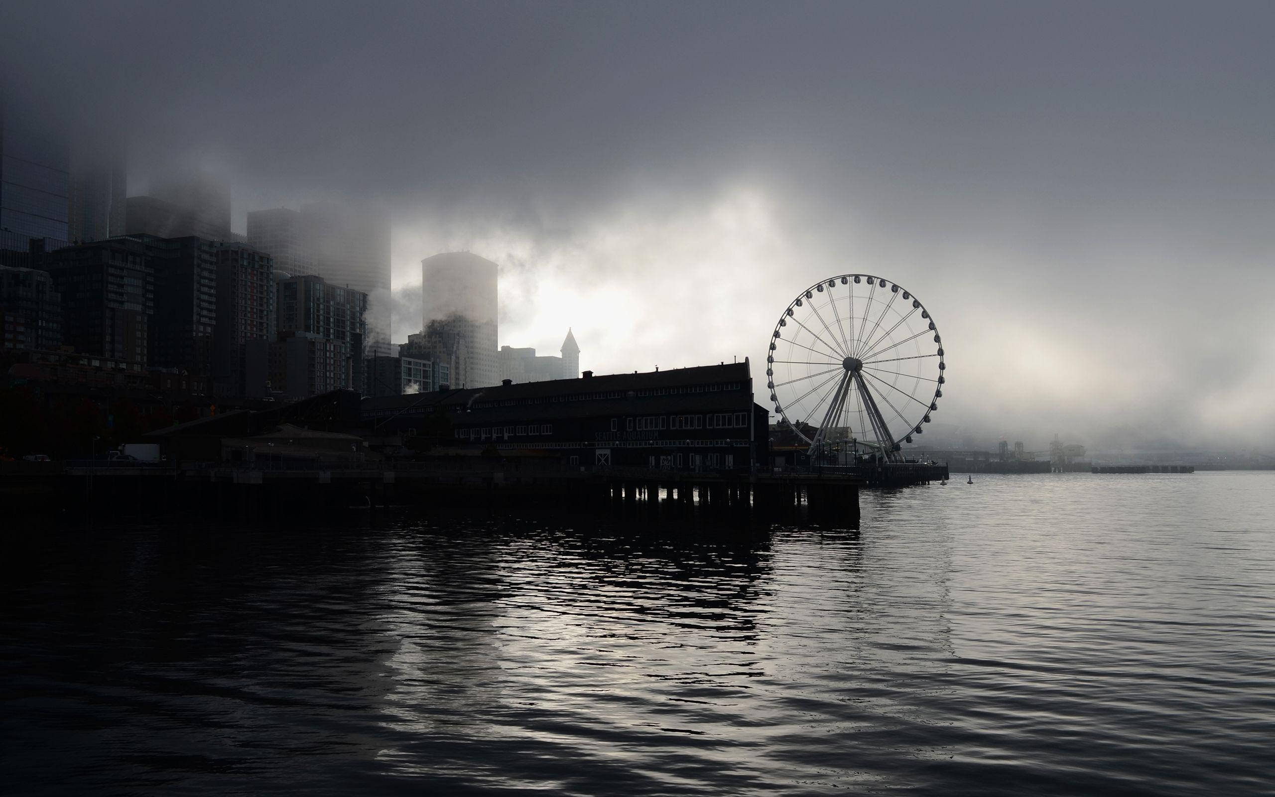 Wheel 桌布 壁紙 — 霧氣瀰漫的海濱景色，摩天輪和城市天際線在陰雲密佈的天空下呈現剪影。