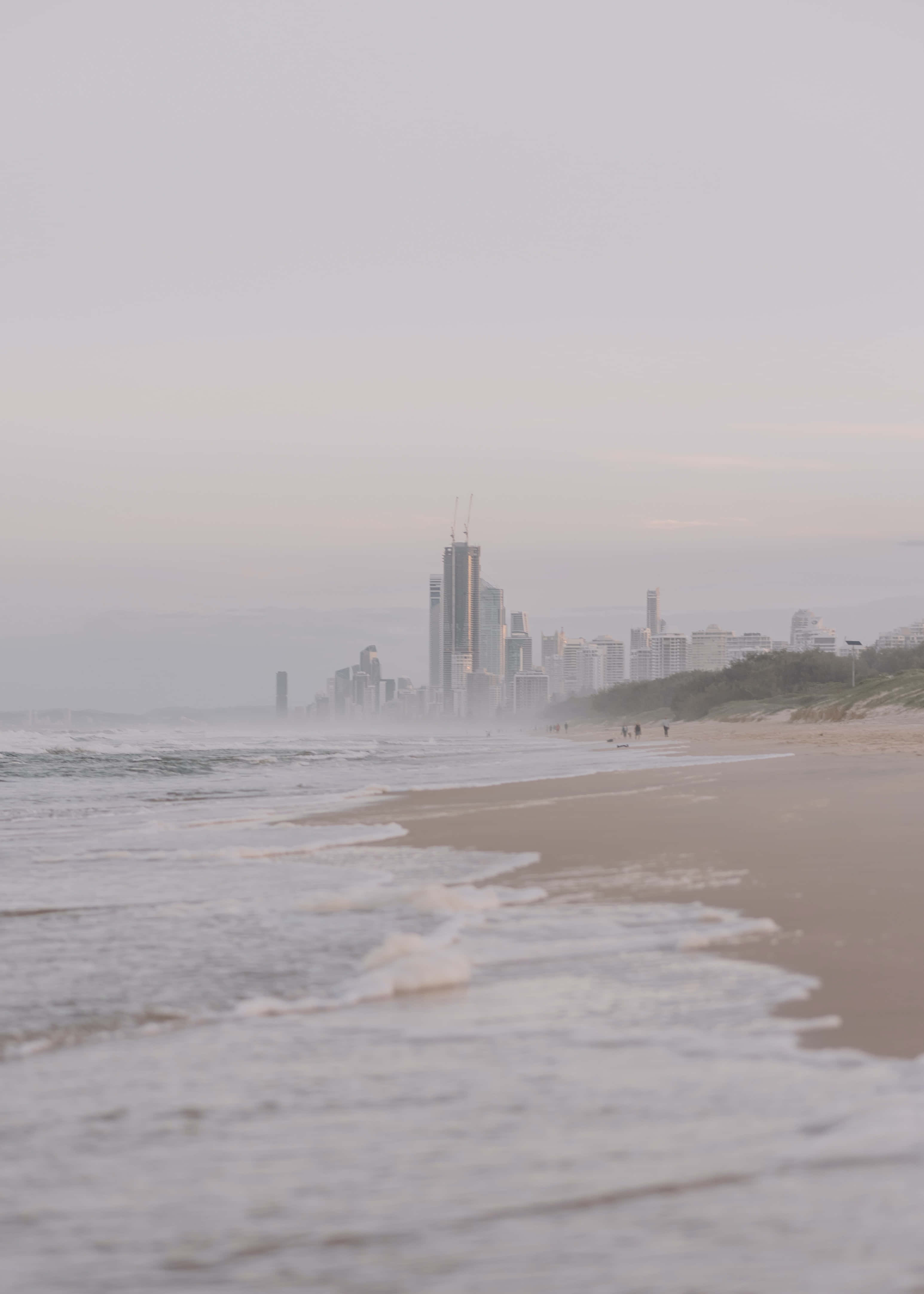 Aesthetic Beachy 桌布 壁紙 — 霧氣瀰漫的海灘，溫柔的海浪輕拍著岸邊，遠處的城市天際線在朦朧的晨光中漸漸消失。