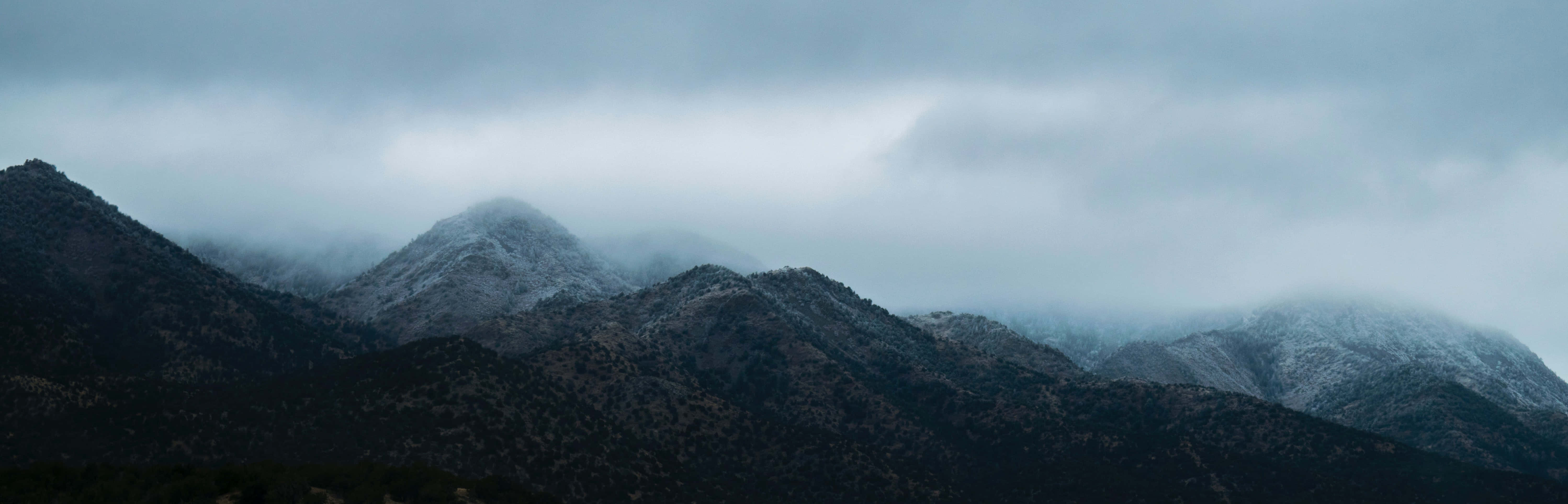 高清 桌布 壁紙 — 霧氣籠罩的山脈，峰頂覆蓋著白雪，在沉重的灰色天空下，呈現出憂鬱的冬季景觀。