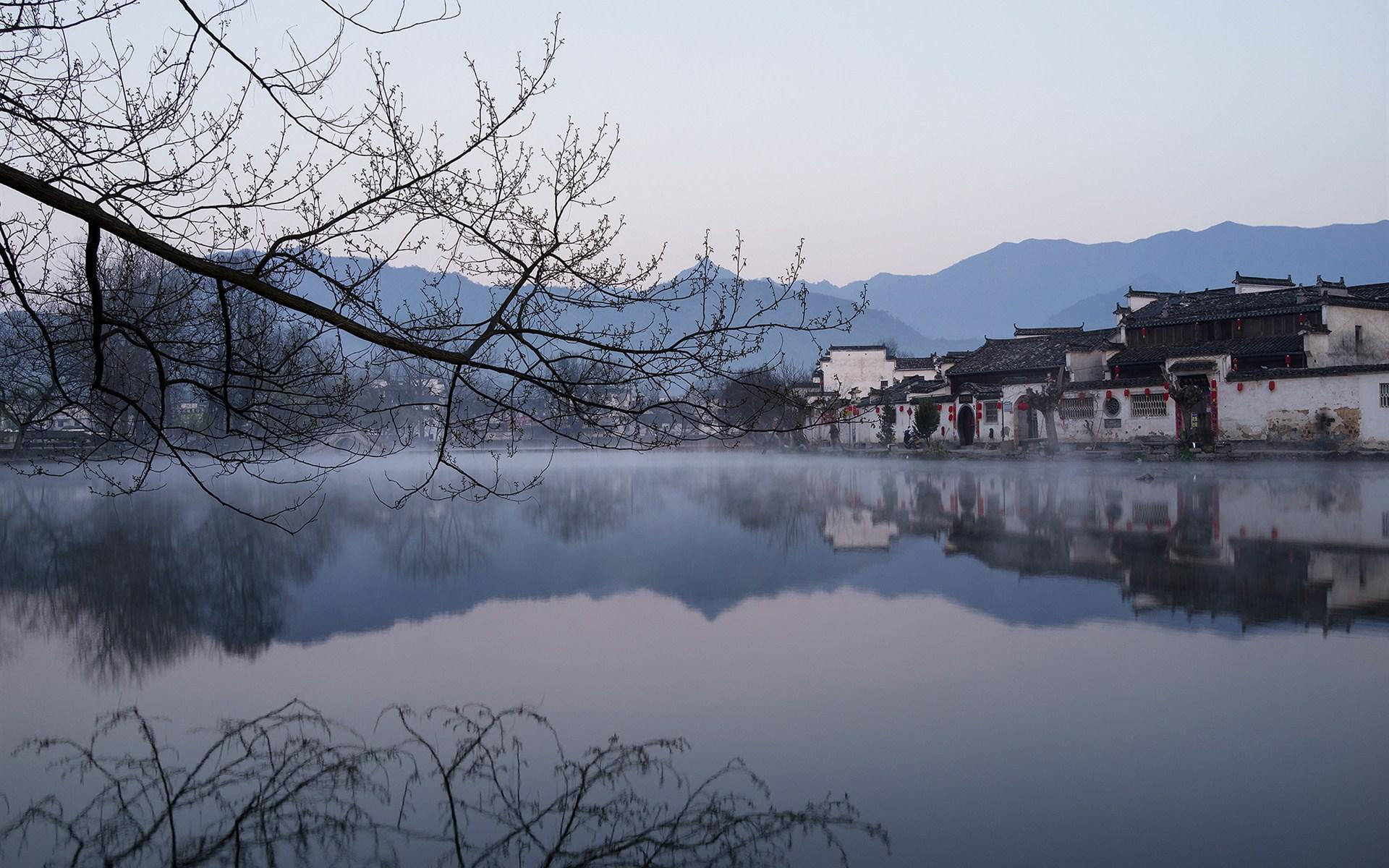 中式房屋壁紙 桌布 壁紙 — 霧氣籠罩的黎明，傳統中國村莊的白牆黑瓦房屋倒映在平靜的水面上，四周環繞著光禿的樹枝。