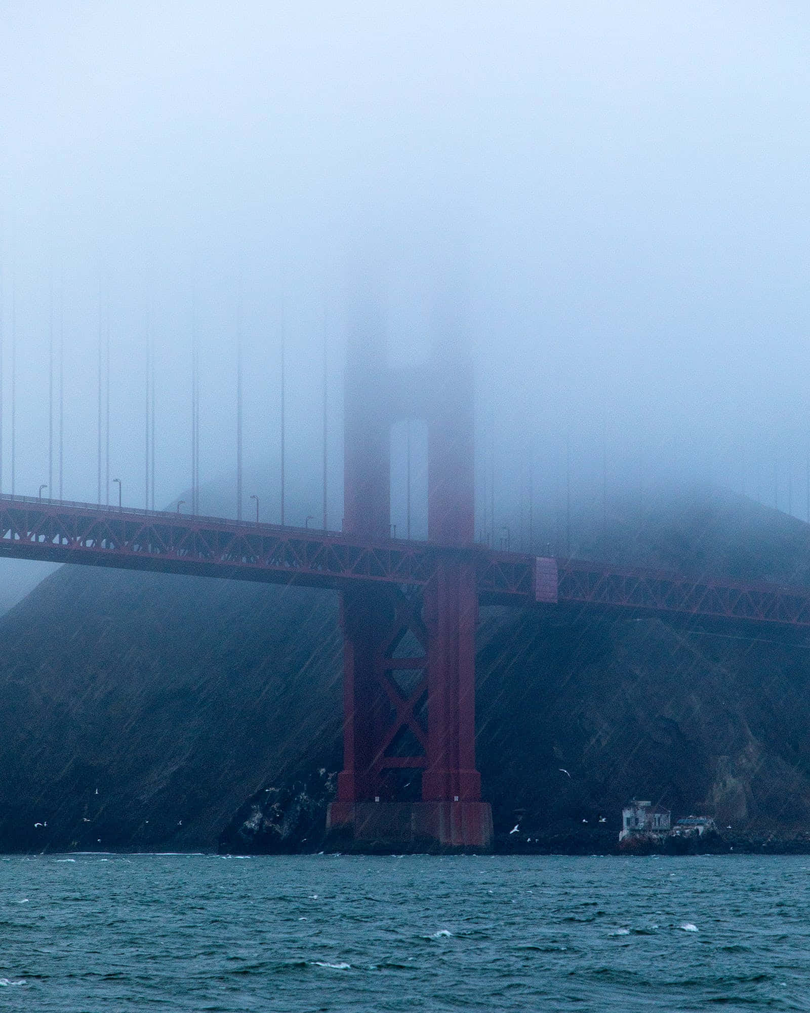 San Francisco Fog 桌布 壁紙 — 霧氣迷霧部分遮擋了標誌性的紅色金門大橋，它橫跨波濤洶湧的海灣，下方是黑暗的懸崖。