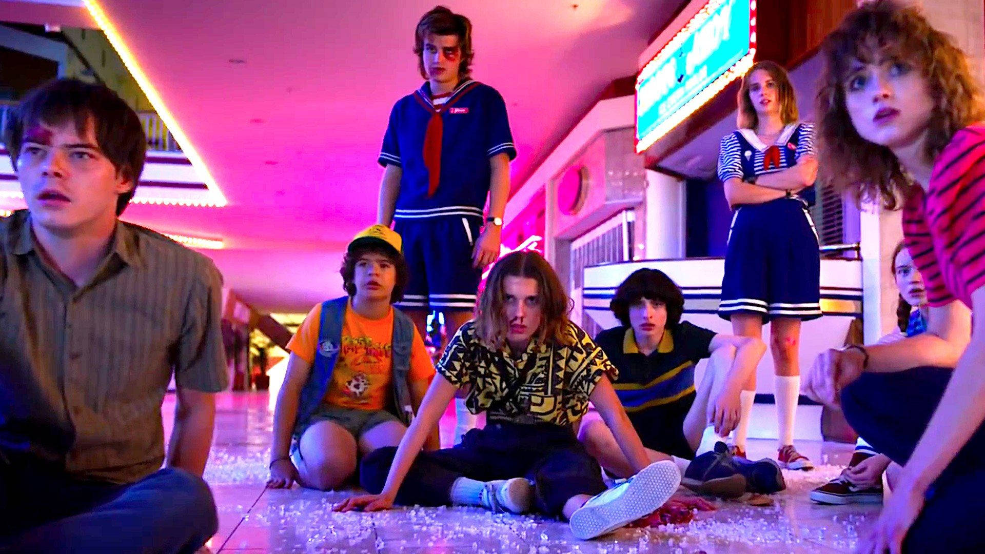 Stranger Things 3 桌布 壁紙 — 青少年穿著1980年代復古服裝，在霓虹燈照亮的遊戲機中心坐著和站著，地上散落著爆米花。