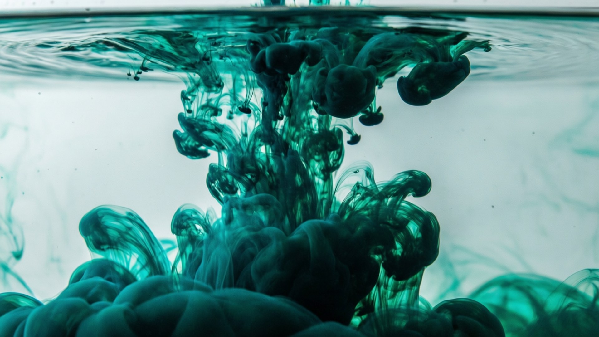 青綠墨水在水中的擴散 壁紙 — 鮮豔的青綠色墨水在清澈的水中下沉時戲劇性地旋轉，形成有機的煙霧狀捲鬚。