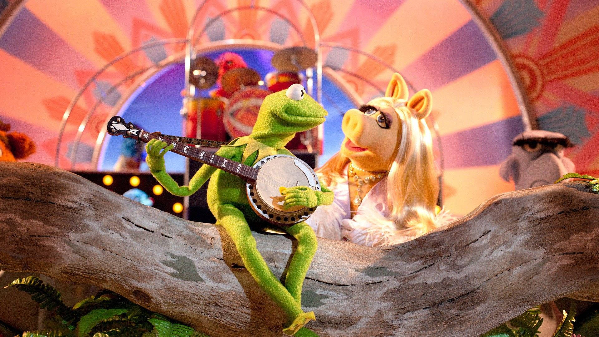 Miss Piggy 桌布 壁紙 — 青蛙柯密特彈奏班卓琴，而豬小姐在他旁邊的木頭上，在色彩繽紛的馬戲帳篷下休息。