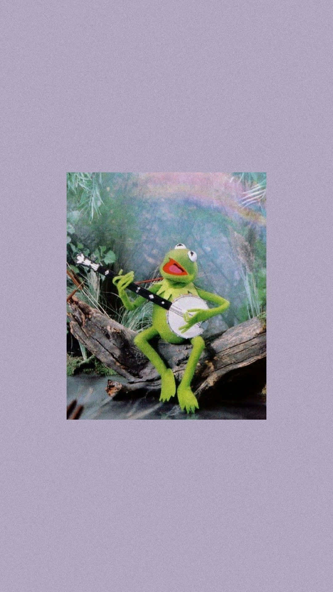 Aesthetic Frog 桌布 壁紙 — 青蛙柯米特在茂密的綠色森林中的一根木頭上彈奏班卓琴，背景有彩虹。