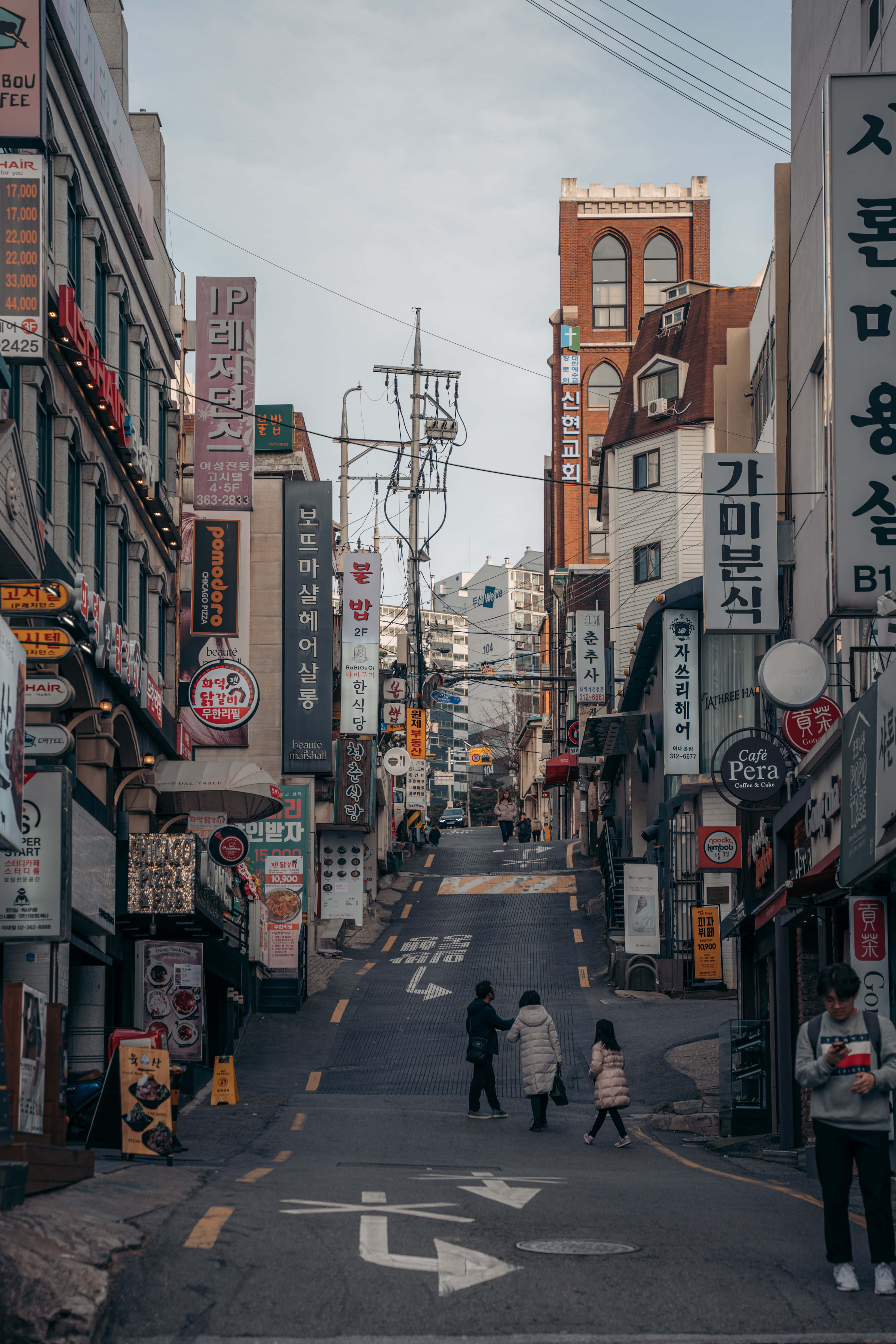 韓國 桌布 壁紙 — 韓國一條狹窄的城市小巷，兩旁掛滿色彩繽紛的招牌，行人在陰天中行走。