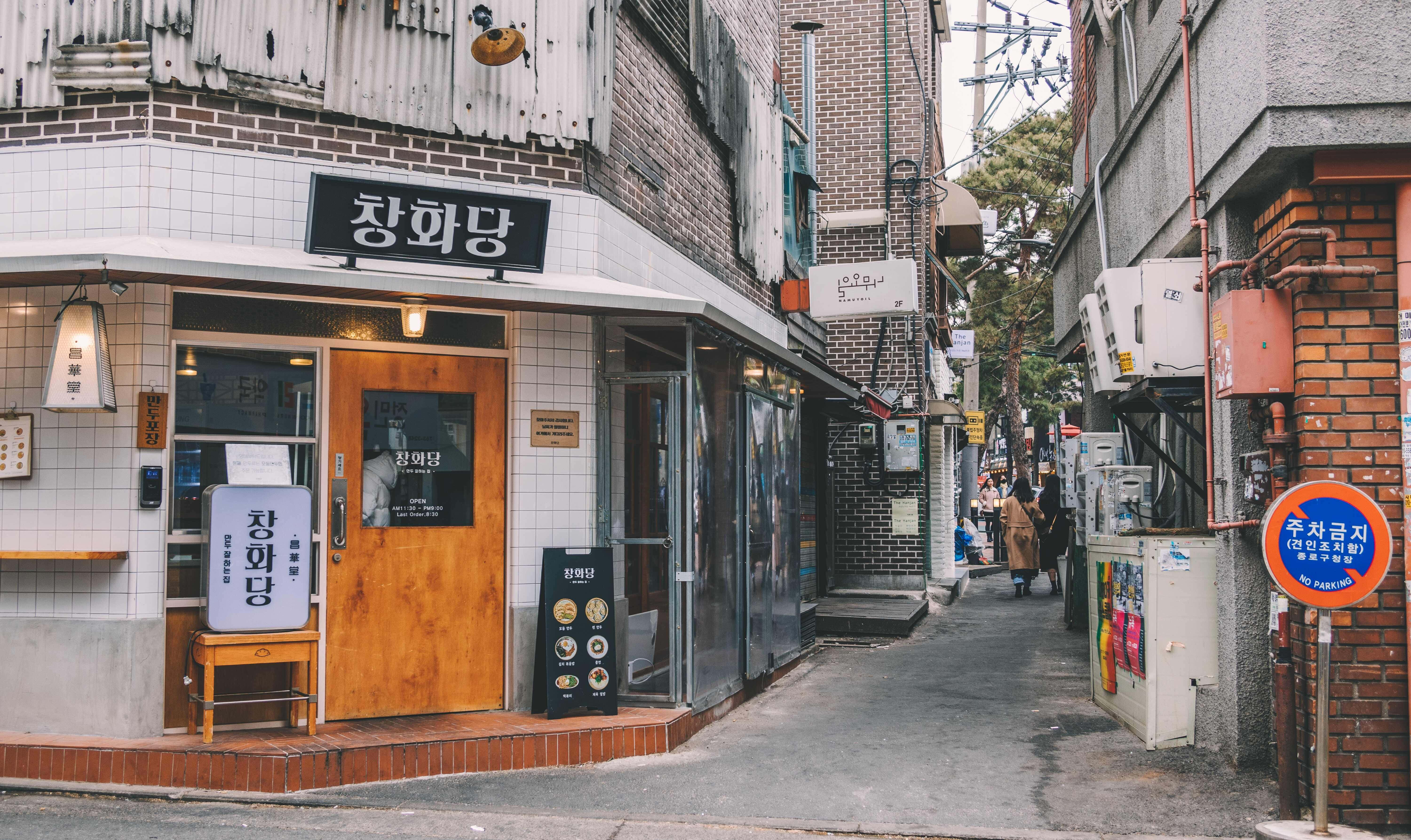 韓國 桌布 壁紙 — 韓國一條狹窄的小巷，兩旁是磚砌建築和一間木門咖啡館，天空多雲。