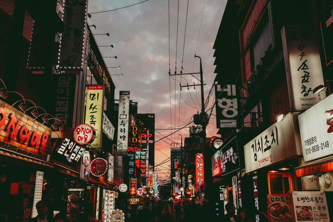 桌布 壁紙 — 韓國城市黃昏時分，一條霓虹燈閃爍的熱鬧小巷，擠滿攤位和發光招牌。