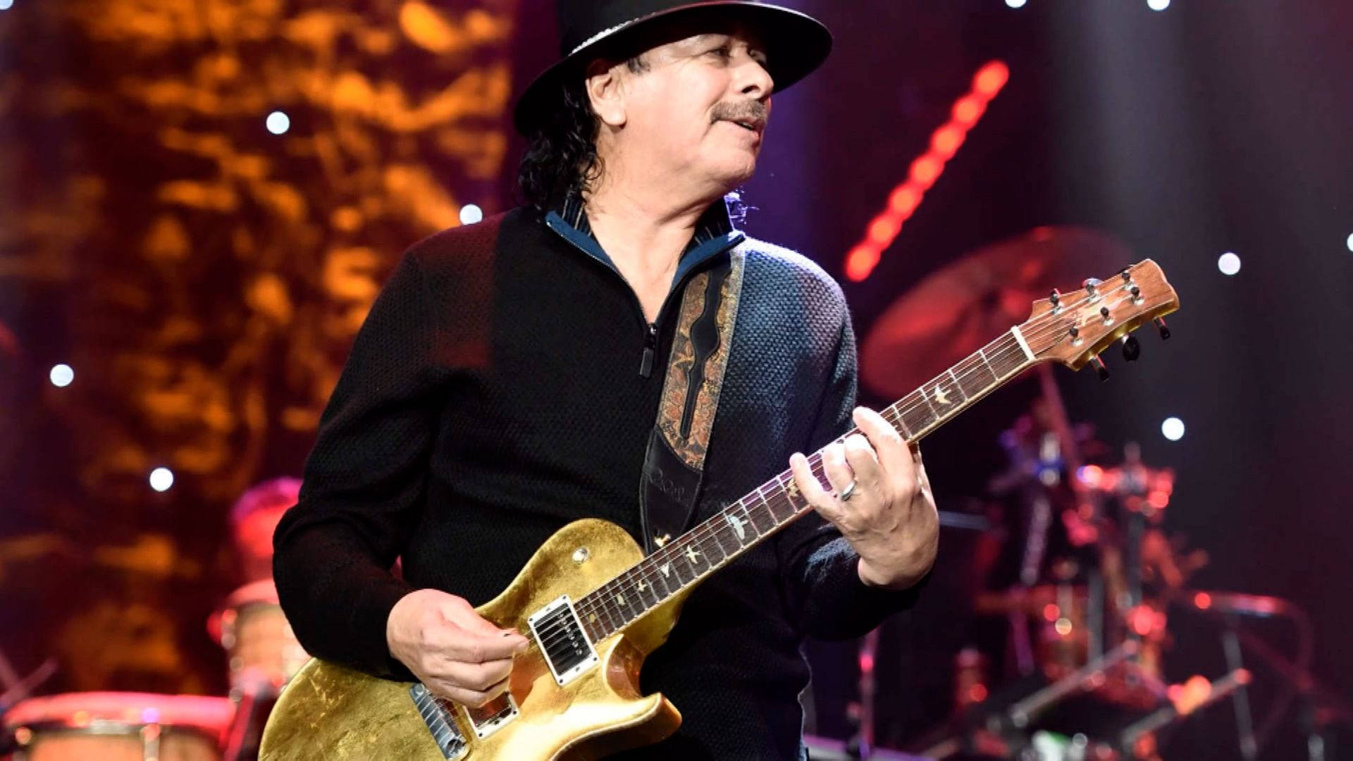 音樂 桌布 壁紙 — 音樂家 Carlos Santana 在溫暖的舞台燈光下，背景有鼓組，演奏金色電吉他。