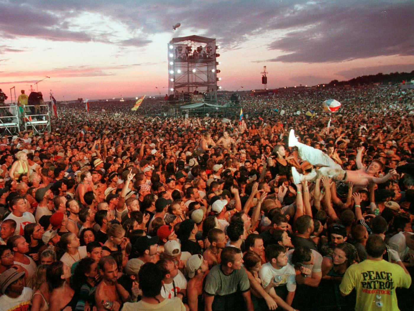 Woodstock 桌布 壁紙 — 音樂節上大量人群在暮色天空下跳舞，遠處有燈光舞台。
