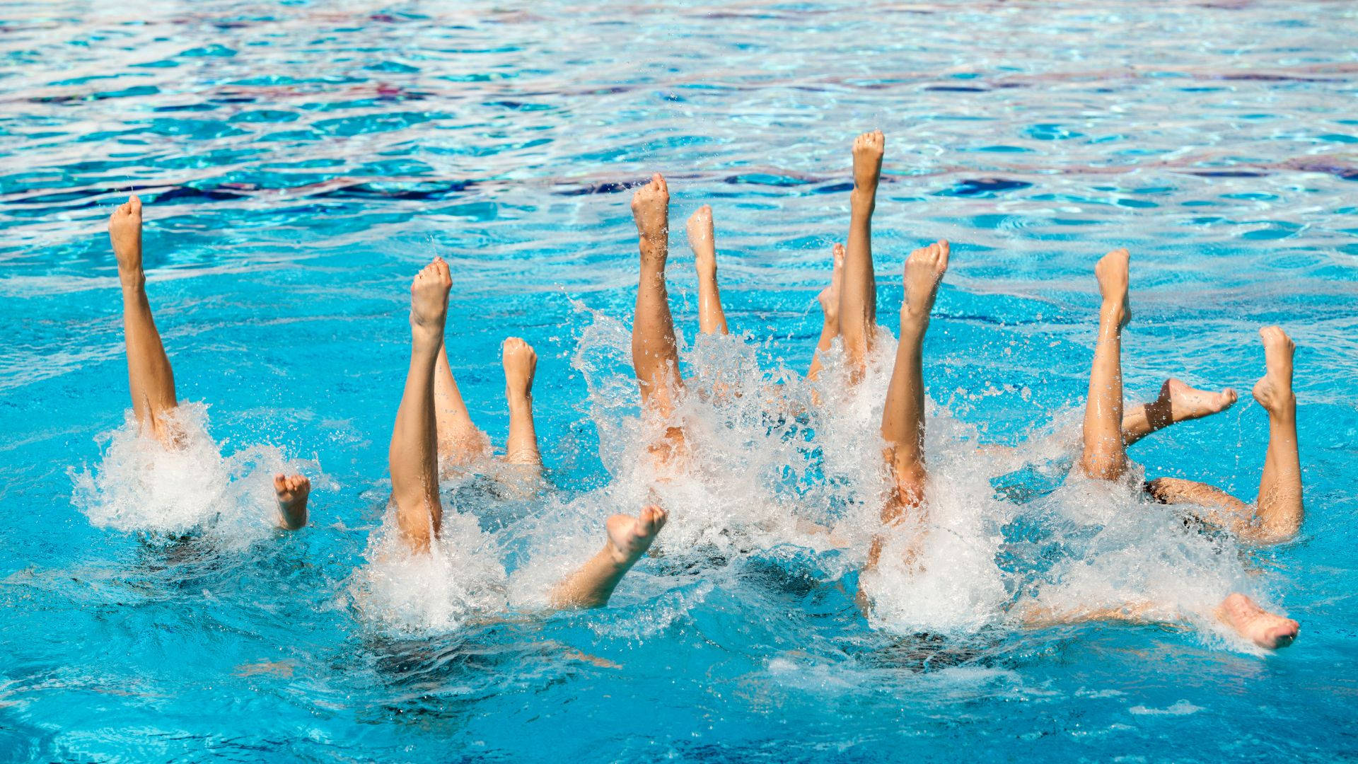Swimming 桌布 壁紙 — 韻律泳選手的雙腿從充滿活力的藍色水中向上射出，形成爆炸性的白色水花。