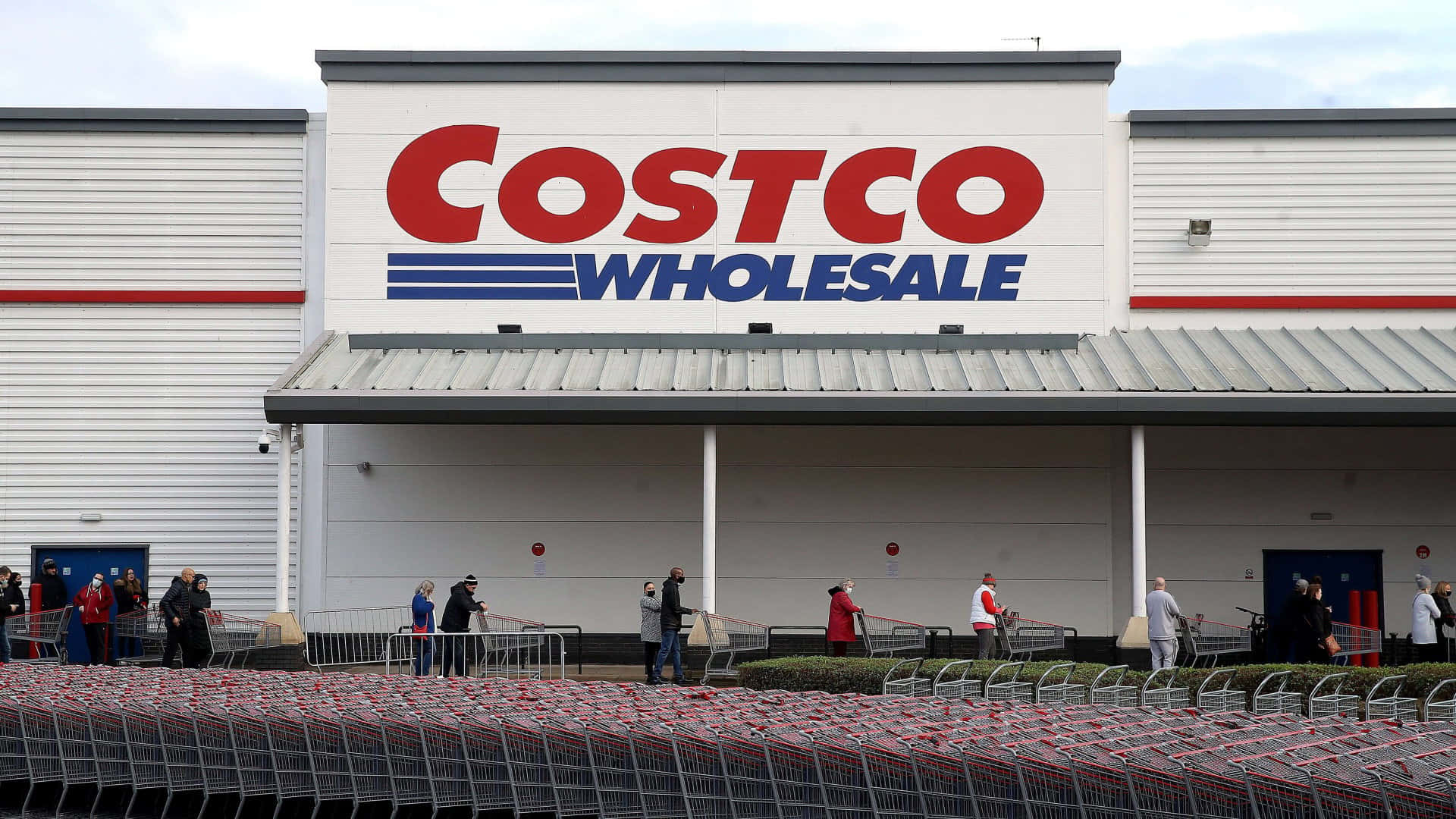 Costco 桌布 壁紙 — 顧客聚集在Costco Wholesale商店外，前面有一排排購物車，天空多雲。