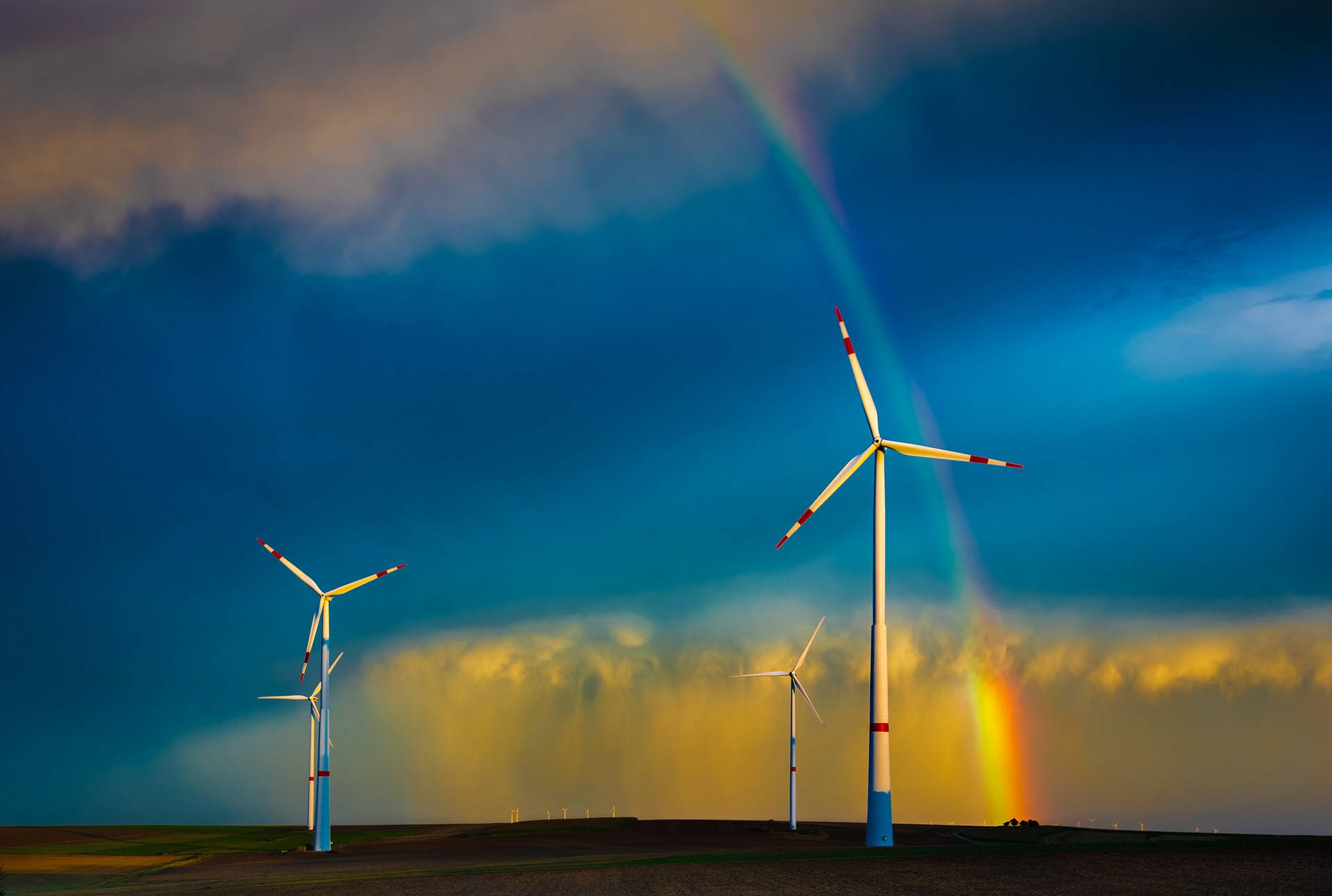 Energy 桌布 壁紙 — 風力渦輪機在戲劇性的天空下高聳，一道鮮豔的彩虹穿過暴風雲，在平坦的田野上彎曲。
