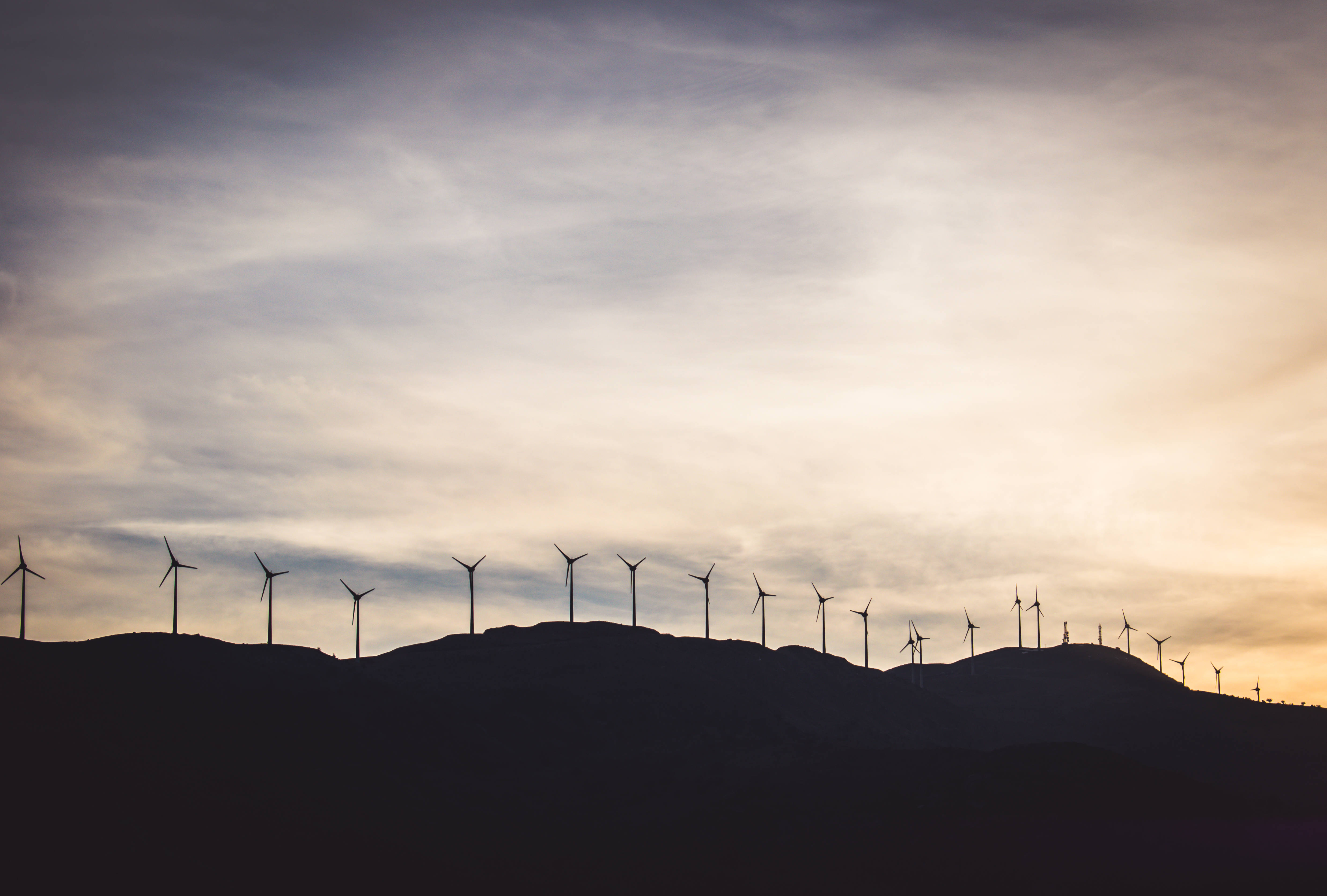 Energy 桌布 壁紙 — 風力發電機的剪影沿著起伏的山丘排列，背景是柔和的金色日落天空，點綴著縷縷薄雲。