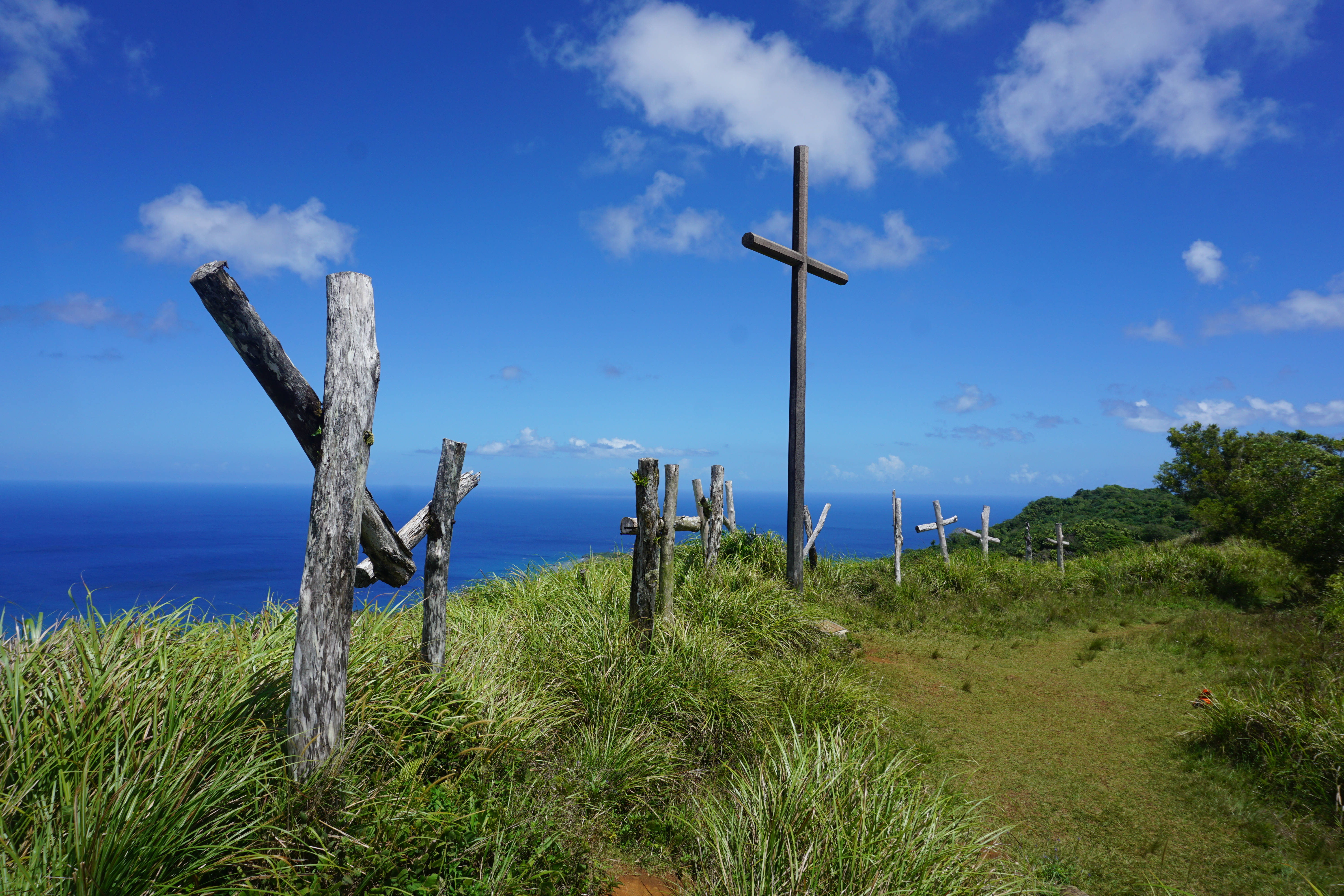 Guam 桌布 壁紙 — 風化的木製十字架矗立在長滿草的懸崖上，俯瞰著廣闊的藍色海洋，天空晴朗，有零星雲朵。
