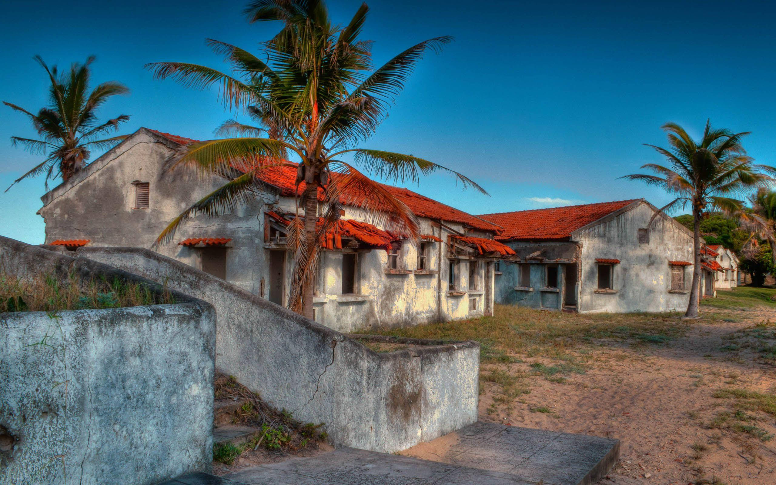 Mozambique 桌布 壁紙 — 風化的白色建築物配紅色瓦片屋頂，在熱帶環境中矗立於鮮豔的藍天下，由高大的棕櫚樹框住。
